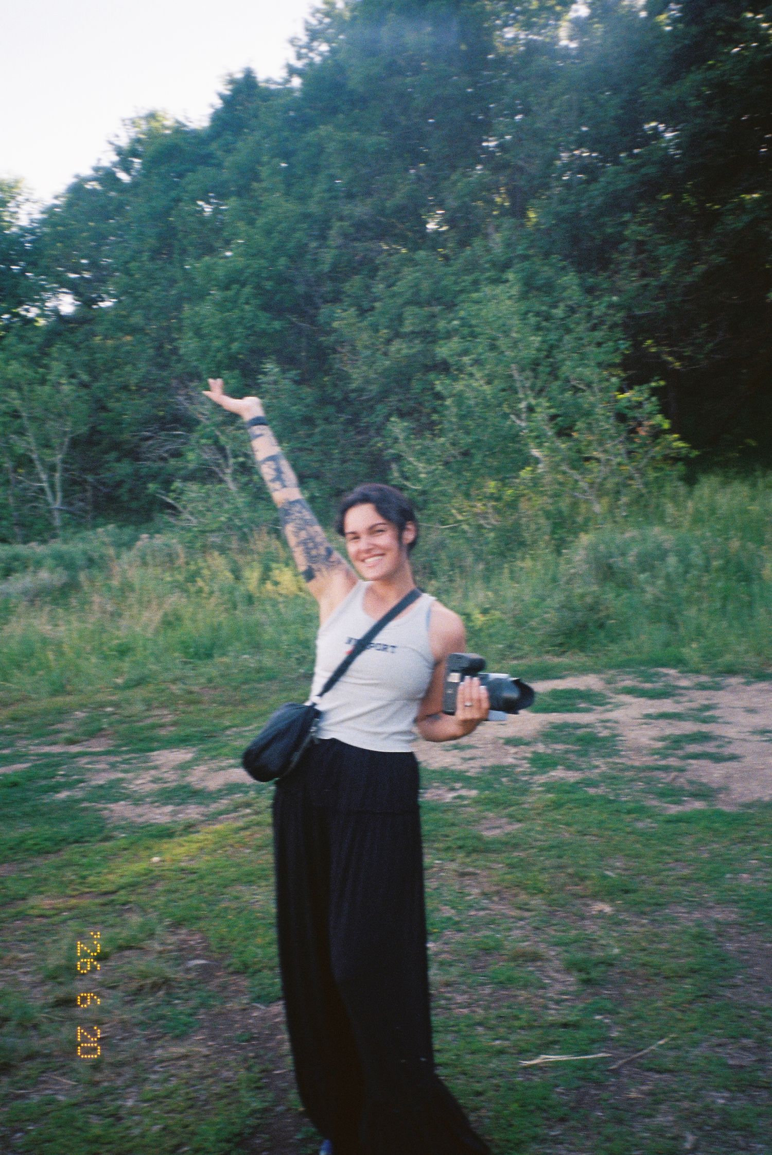 Smiling woman with a tattooed arm raised high holds a camera while standing in a sunlit grassy clearing surrounded by green trees.
