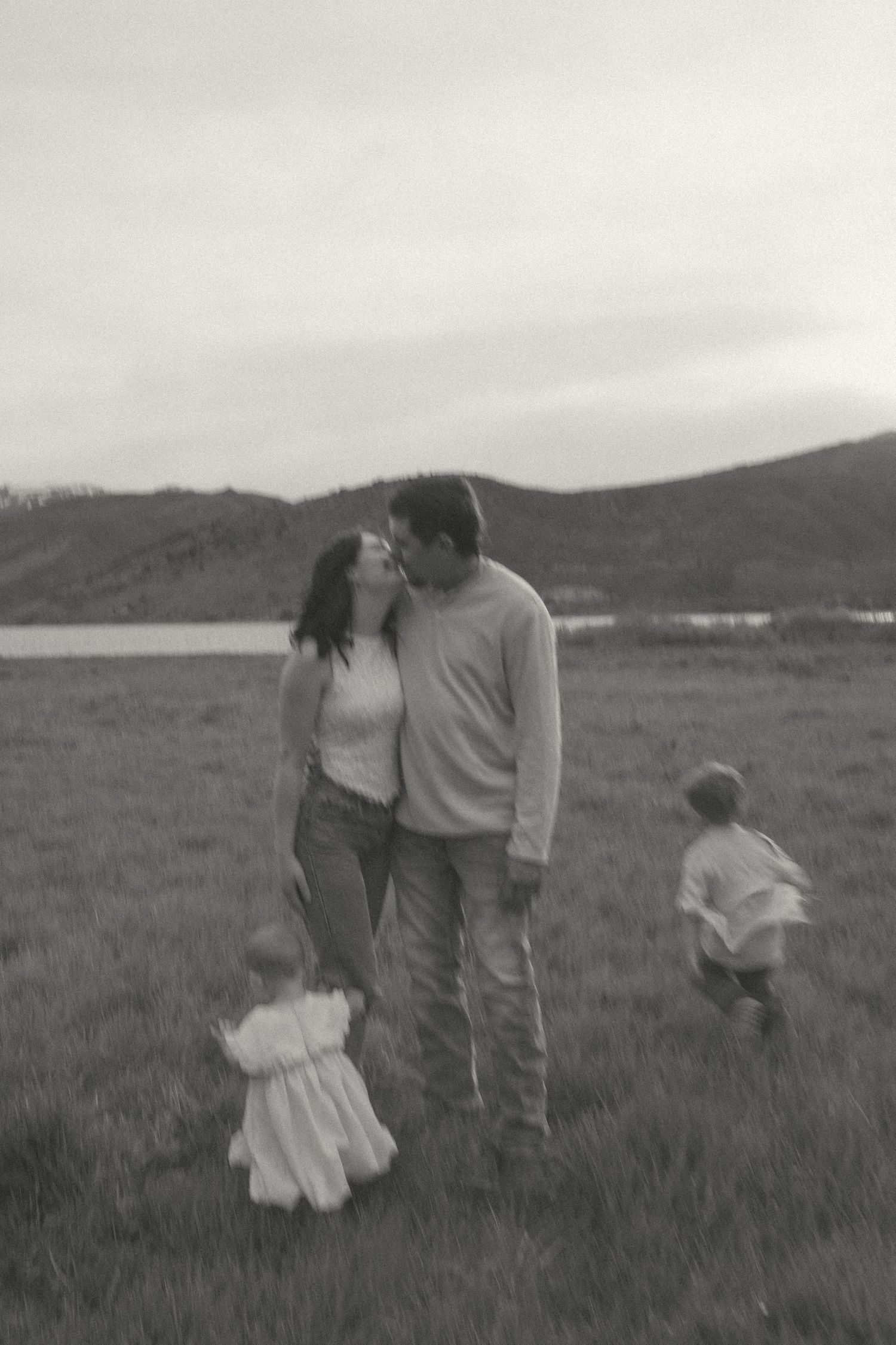 Black and white photo of a couple kissing in a grassy field with two young children playing nearby and hills and a lake in the background.