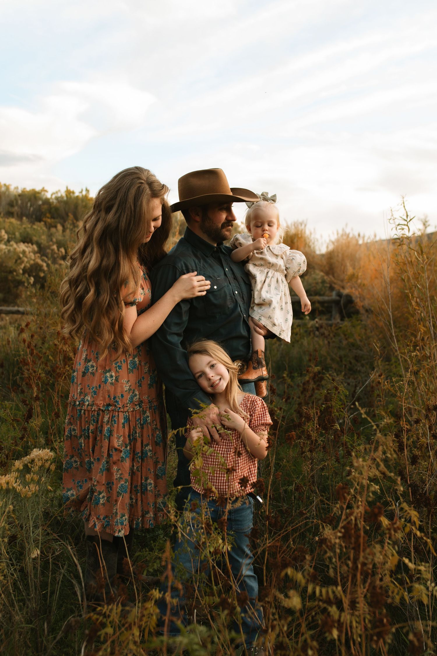 Family of four in coordinated boho outfits posing together in a golden meadow at sunset, with a father in a cowboy hat holding a baby.