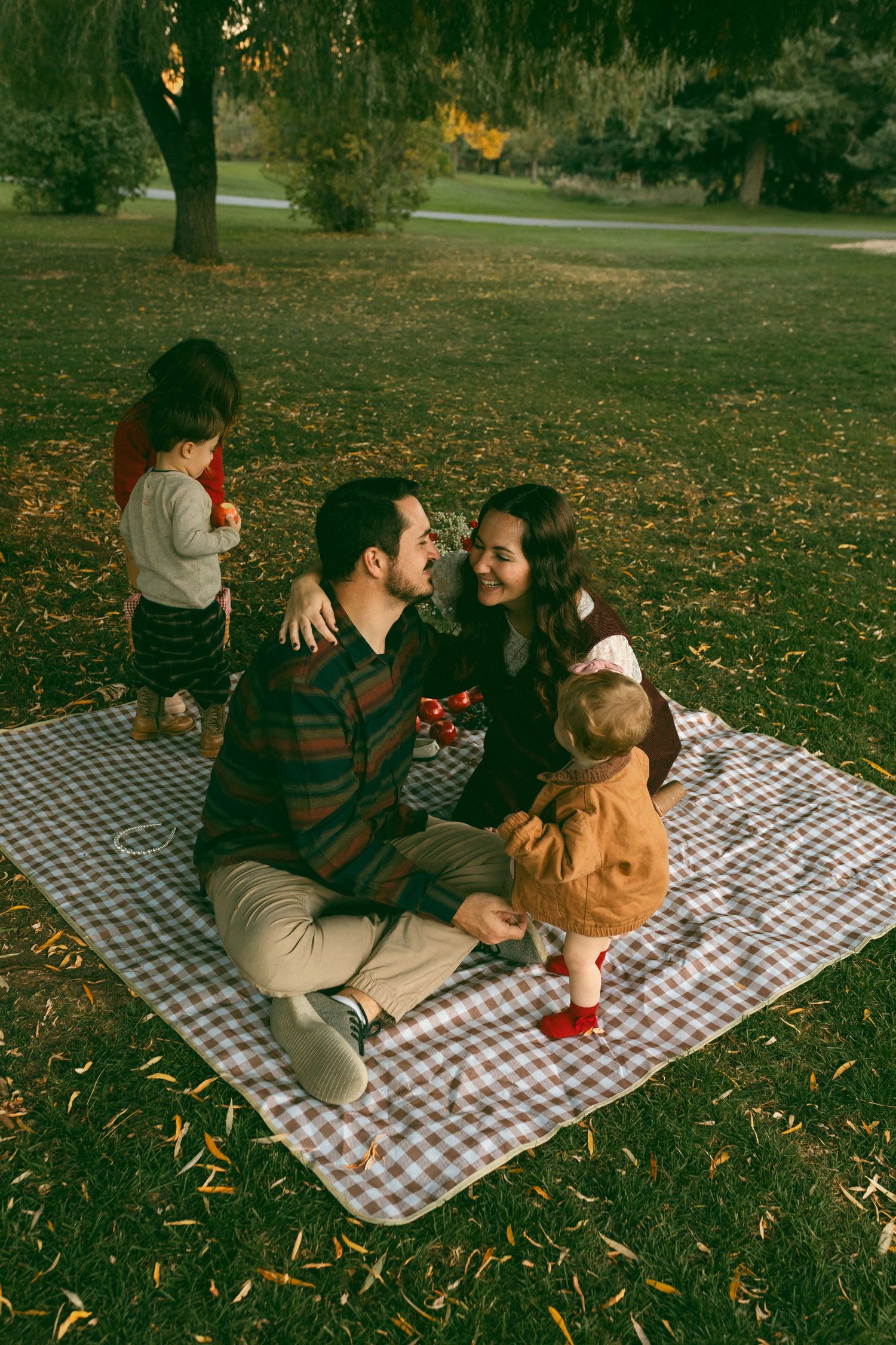 A smiling couple sits close on a checkered picnic blanket in a sunlit park with two toddlers, surrounded by autumn leaves.
