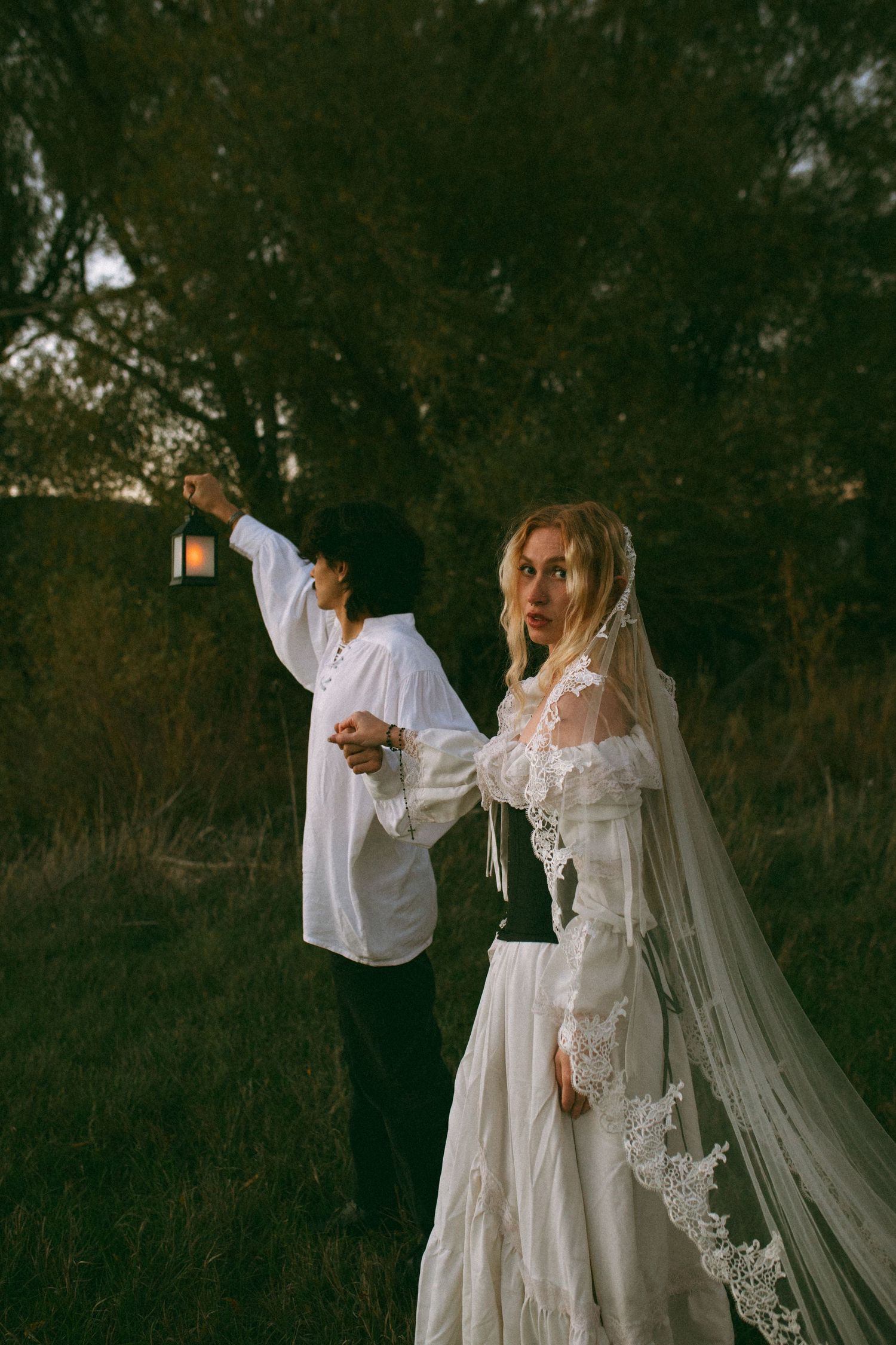 Bride in a lace gown and long veil stands in a grassy field at dusk while a companion in a white shirt holds up a lantern.