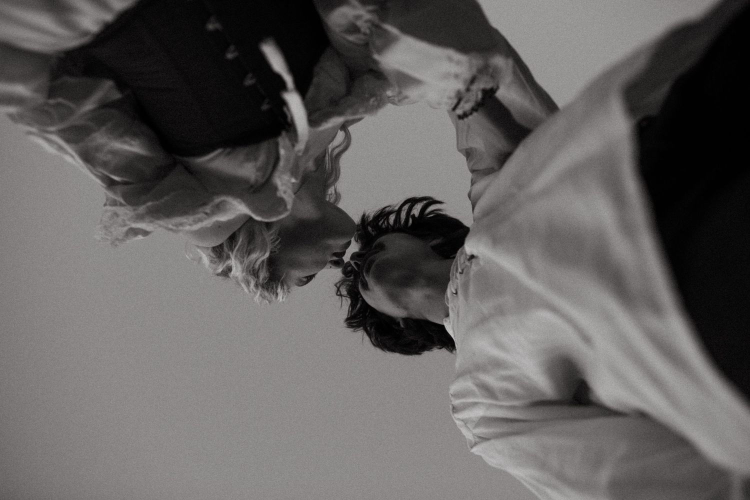 Black and white upside-down view of two people in flowing white clothing dancing closely together against a plain pale sky.