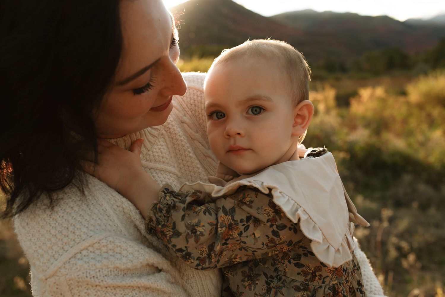 A smiling woman with dark hair holds a curious baby in a floral outfit outdoors, with golden sunset light and hills behind them.
