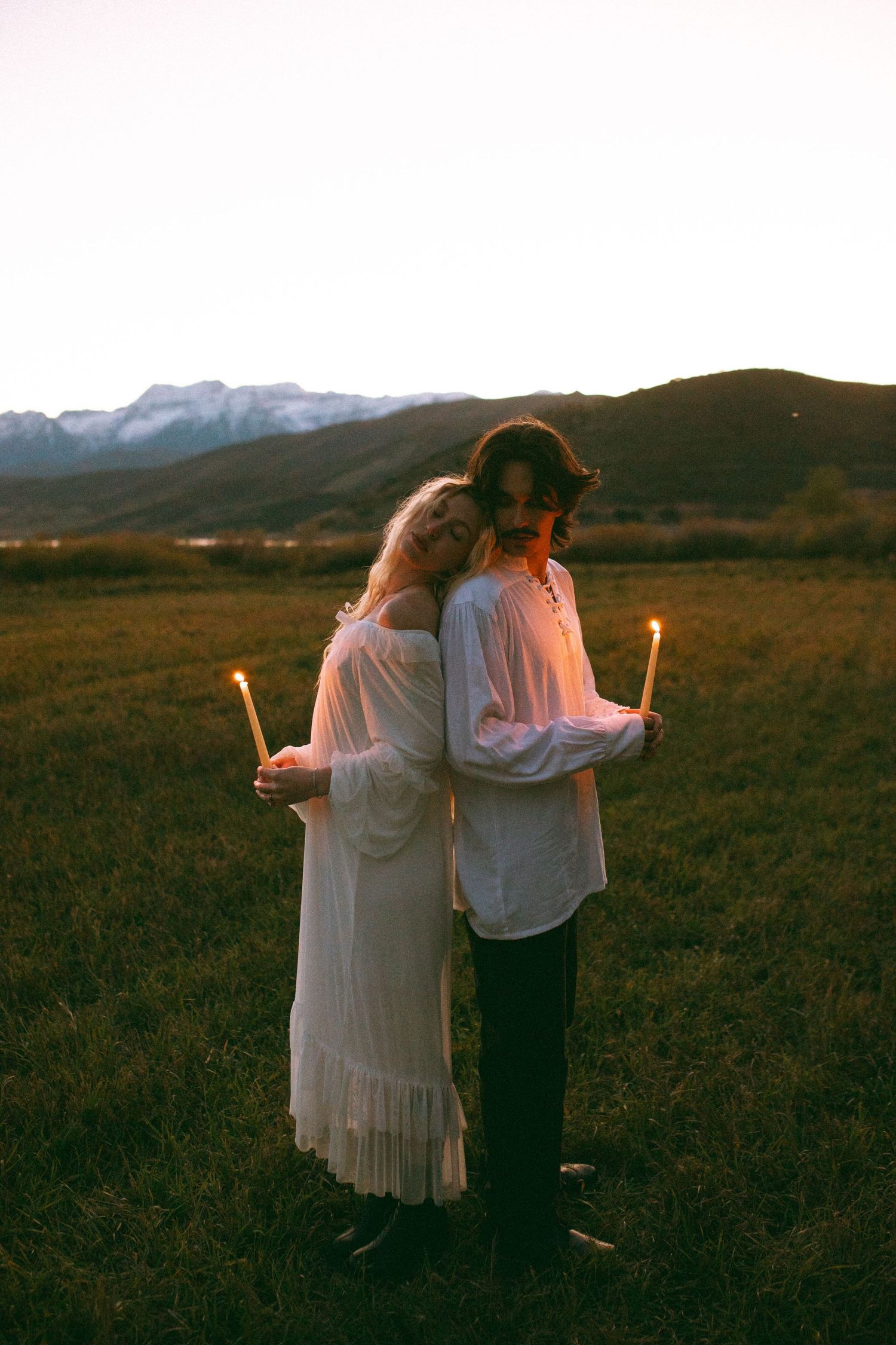 Couple standing back to back holding lit candles in a grassy field at dusk with snowy mountains in the background.