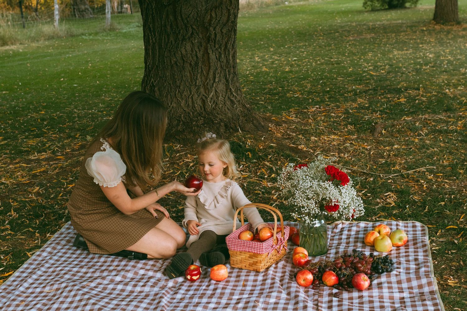 A mother and young daughter enjoy an autumn picnic on a checkered blanket under a large tree, with apples, a basket and red flowers.