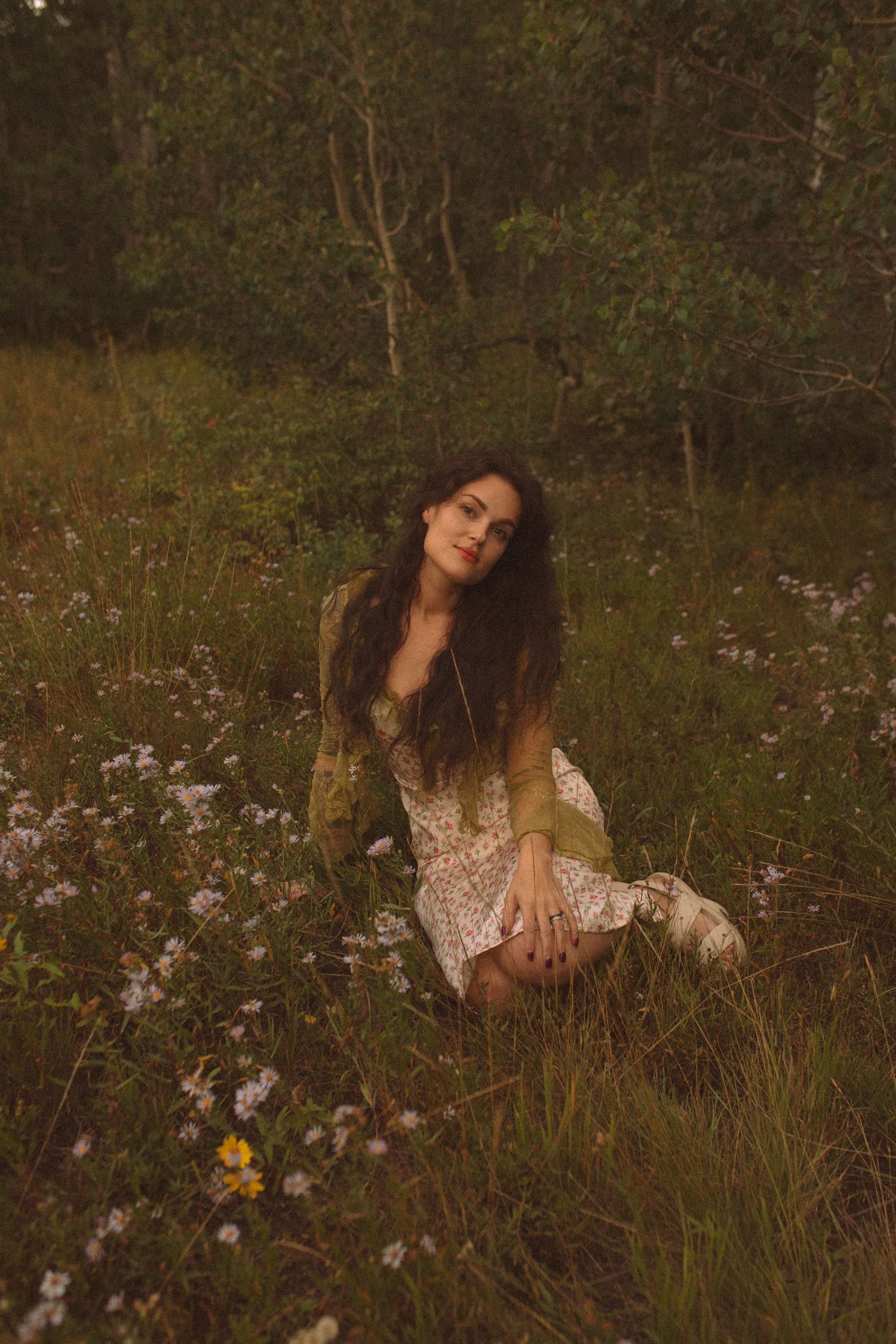Woman with long dark wavy hair sits in a meadow of wildflowers wearing a floral dress and green cardigan, with trees behind her at dusk.