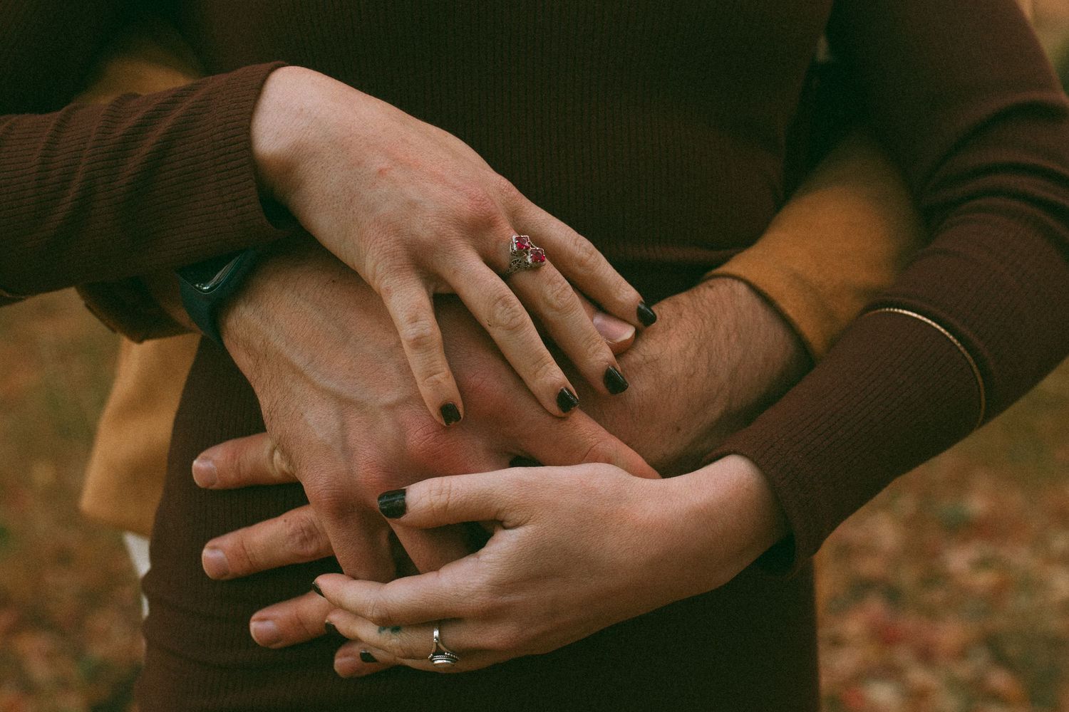 Close-up of a couple's hands clasped together over a torso, with rings and dark nail polish, in warm autumn tones with blurred fall leaves.