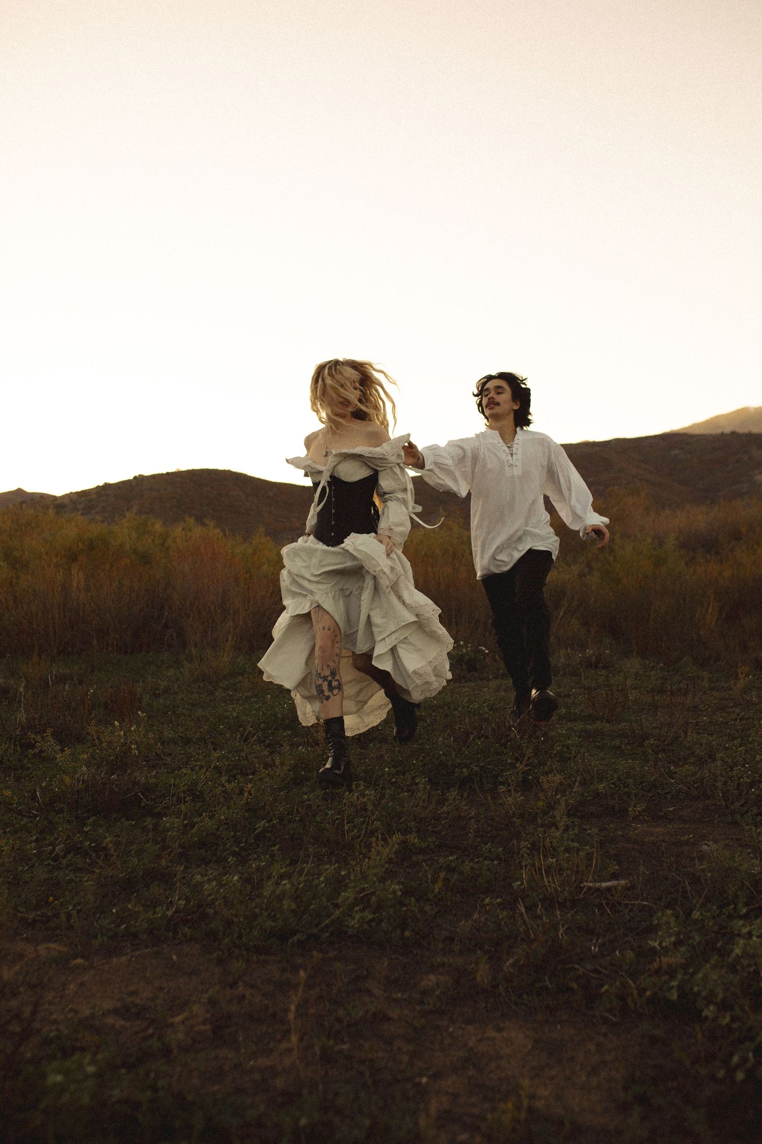 Two people in vintage-style clothing, one in a corset and flowing skirt, run hand in hand through a grassy field at golden hour.