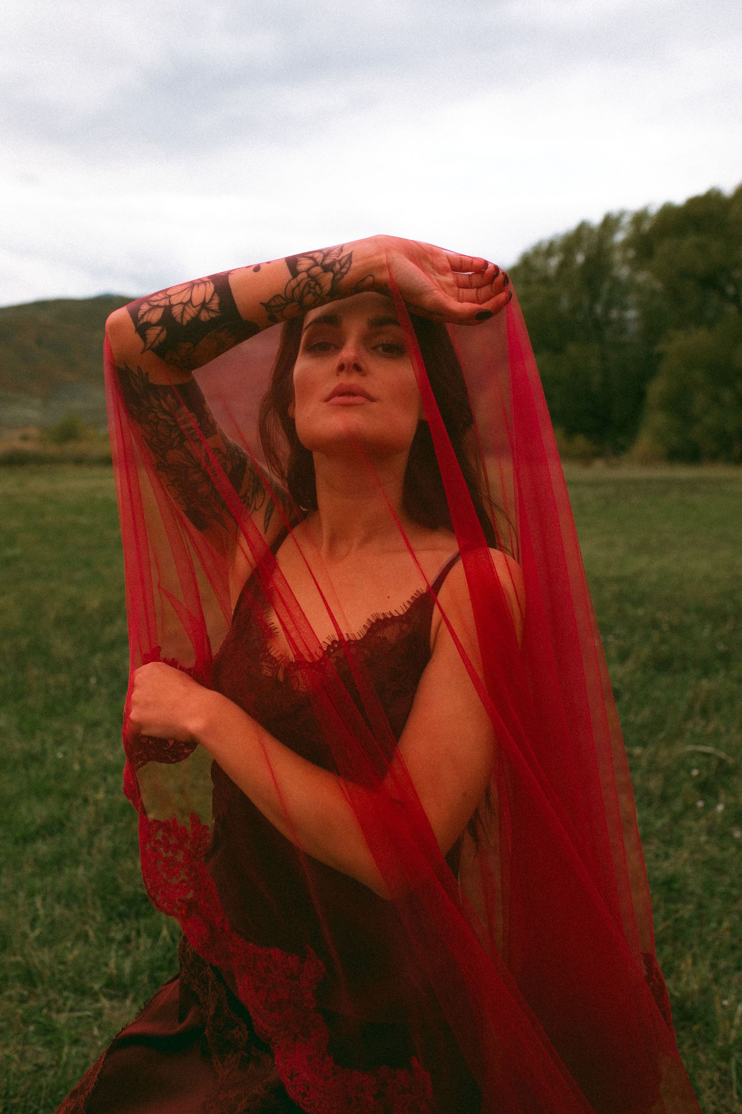 Woman with tattooed arms raised overhead, draped in a sheer red veil and lace slip dress, posing in a grassy field beneath a cloudy sky.