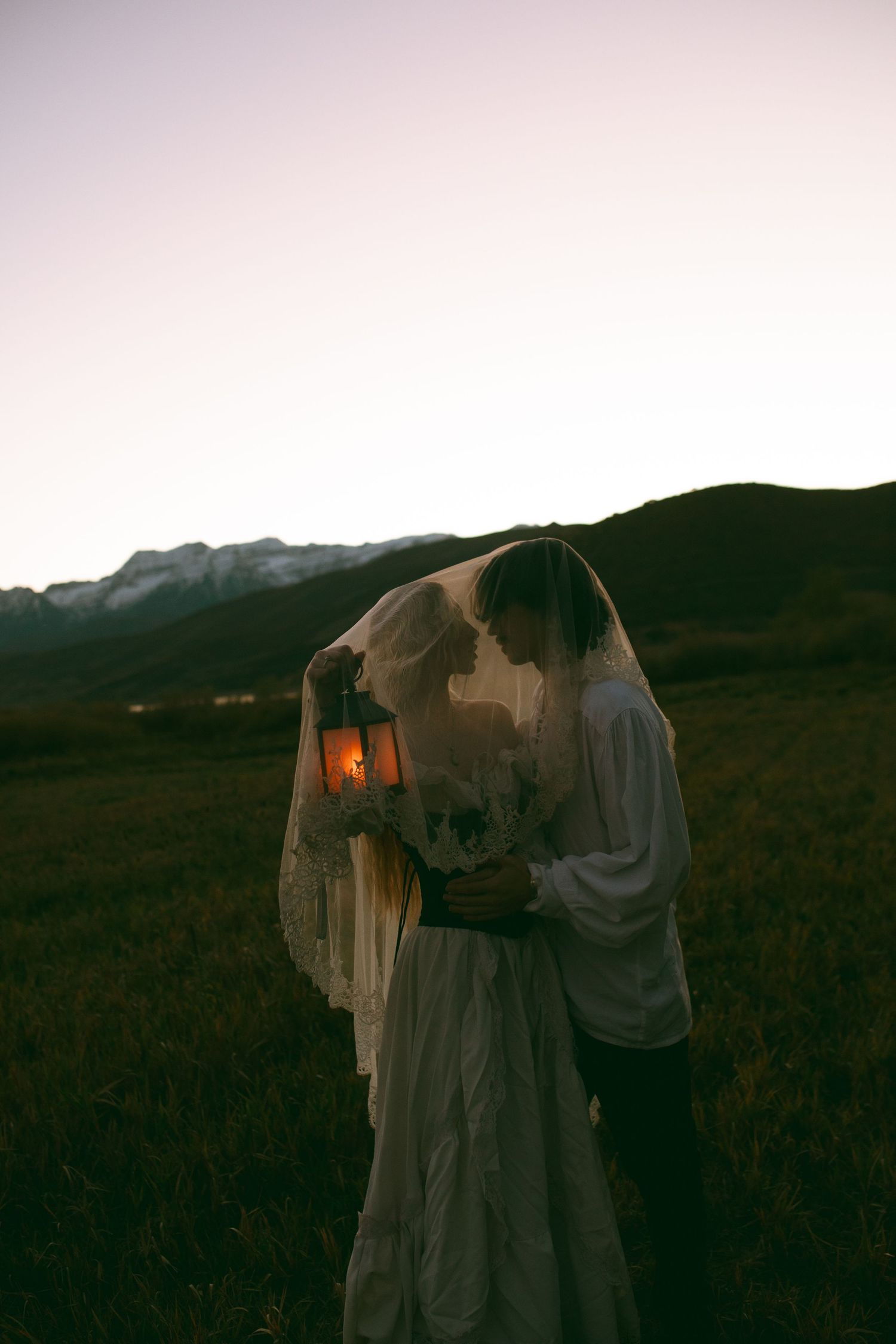 Couple embracing under a lace veil at dusk, holding a glowing lantern, with grassy hills and snowy mountains in the background.