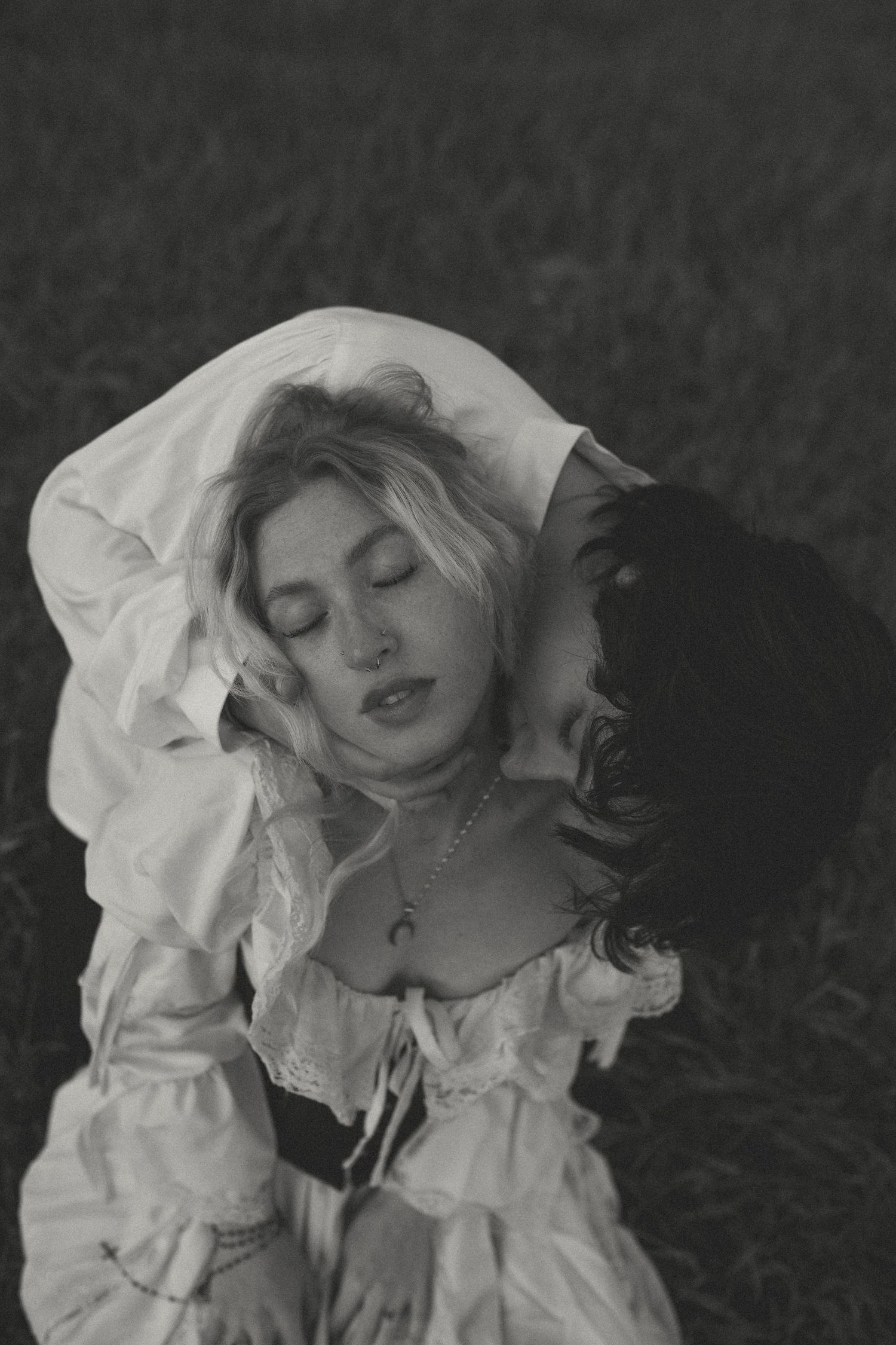 Black and white portrait of a woman with closed eyes in a lace-trimmed white blouse, embraced by a dark-haired person in a grassy field.