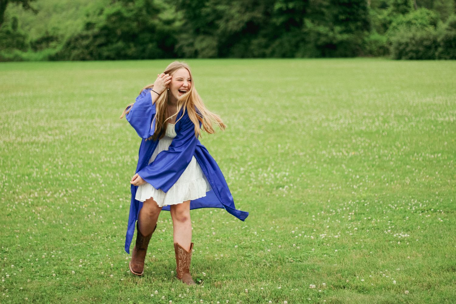 Joyful graduate in a blue gown and cowboy boots laughing in an open field, celebrating a milestone with pure happiness