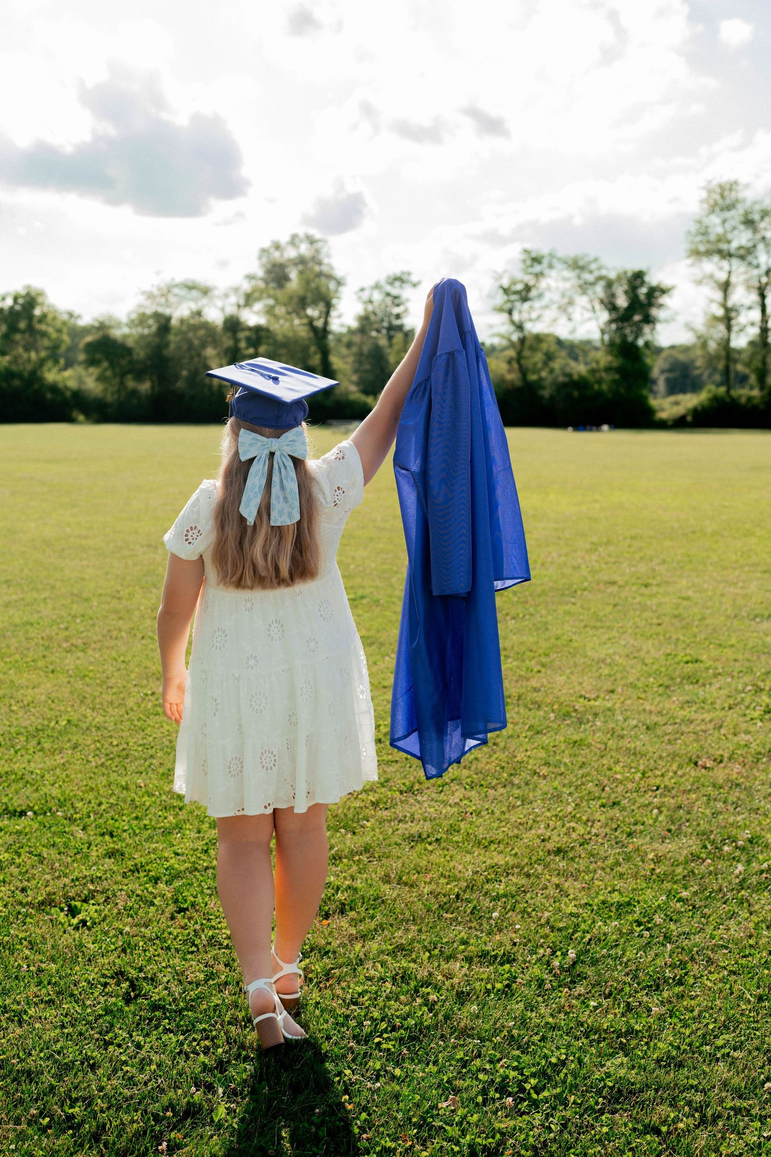 Graduation celebration photo of a girl walking across a sunlit field, holding her blue gown high, symbolizing achievement and new beginnings