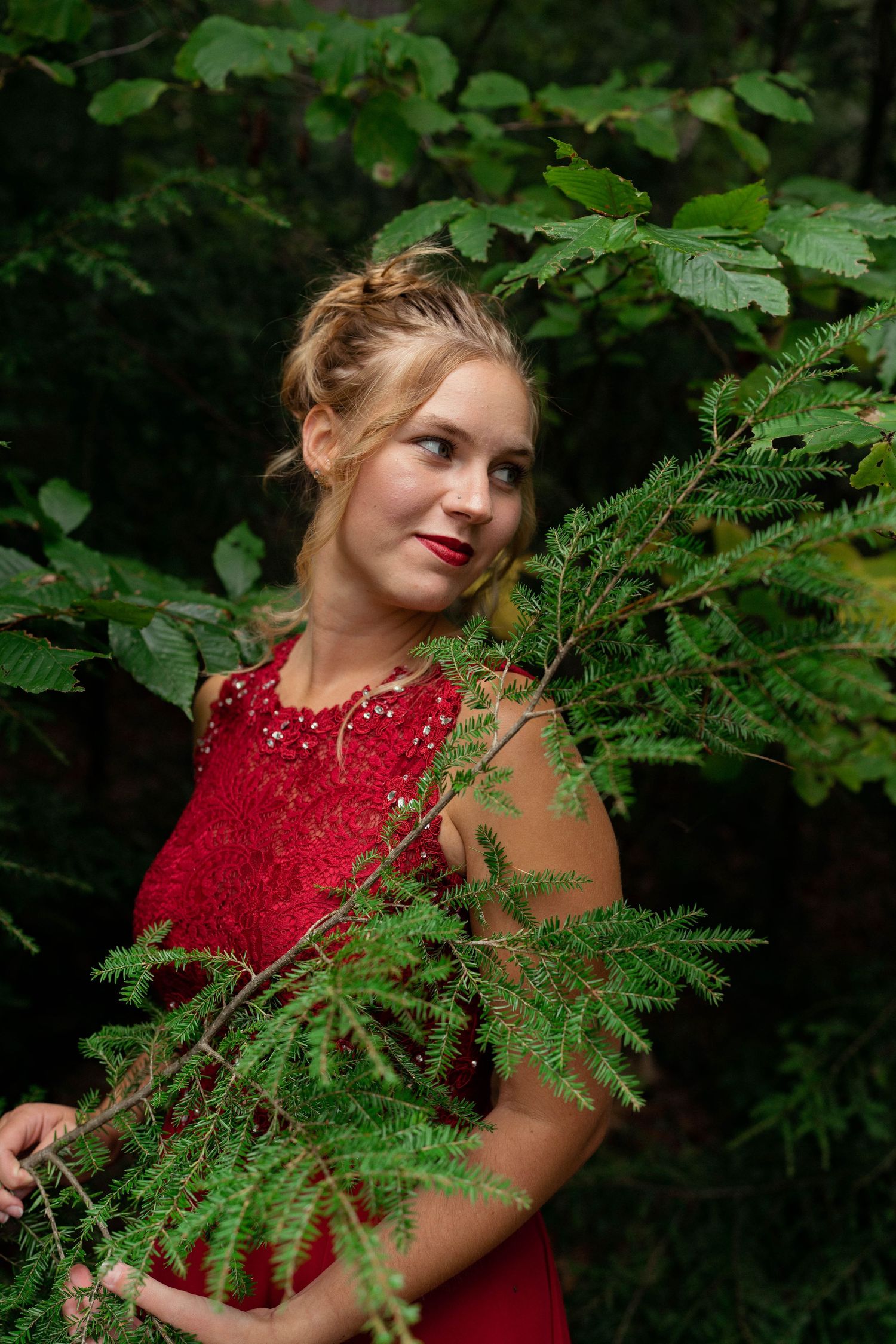 Elegant woman in a red lace dress posing in a lush green forest, gazing to the side. Outdoor portrait photography with natural beauty