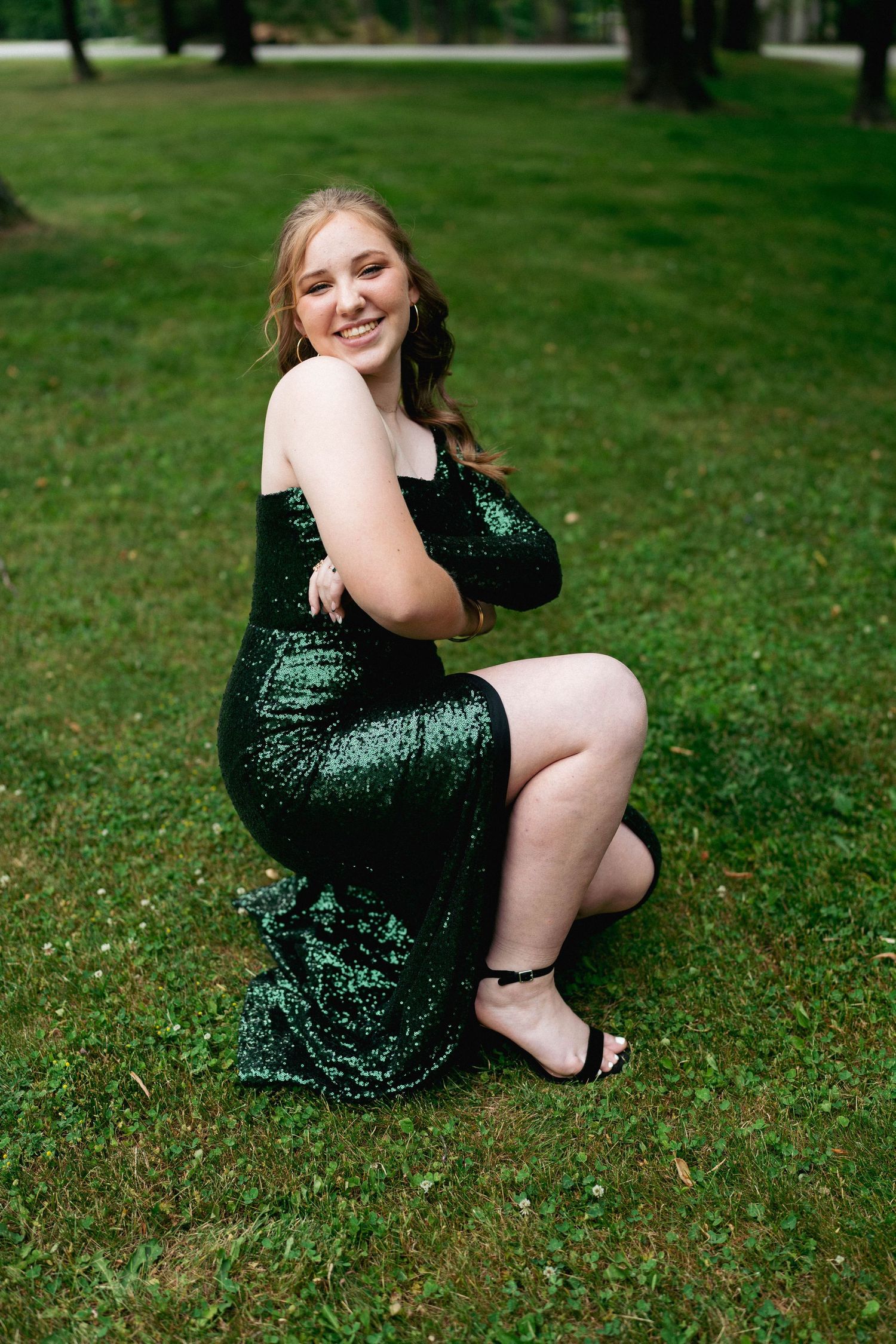 Young woman in an elegant green sequin gown poses confidently on a lush lawn, radiating joy and sophistication
