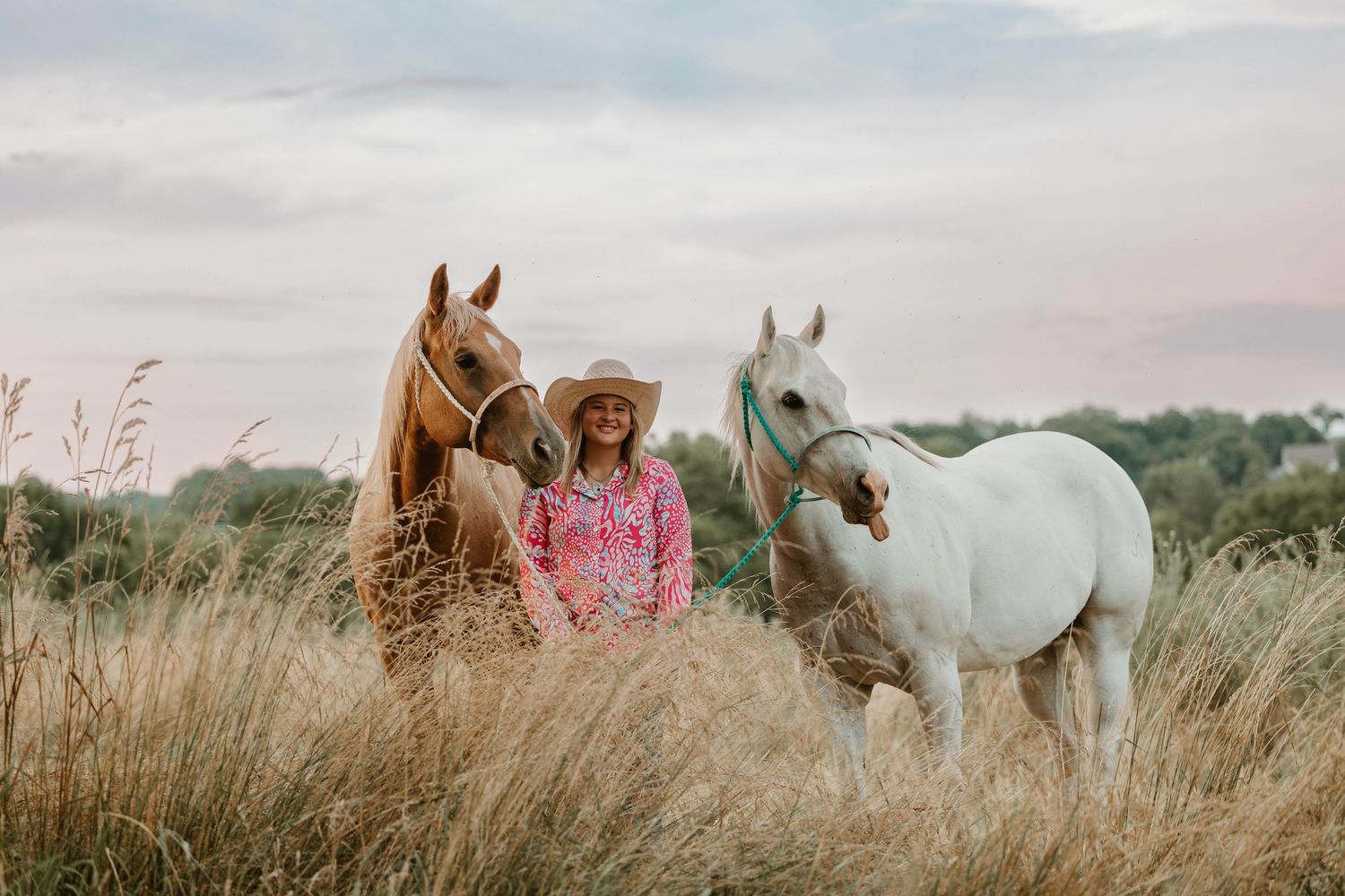 Young girl in cowboy hat stands between brown and white horses in golden field at sunset.