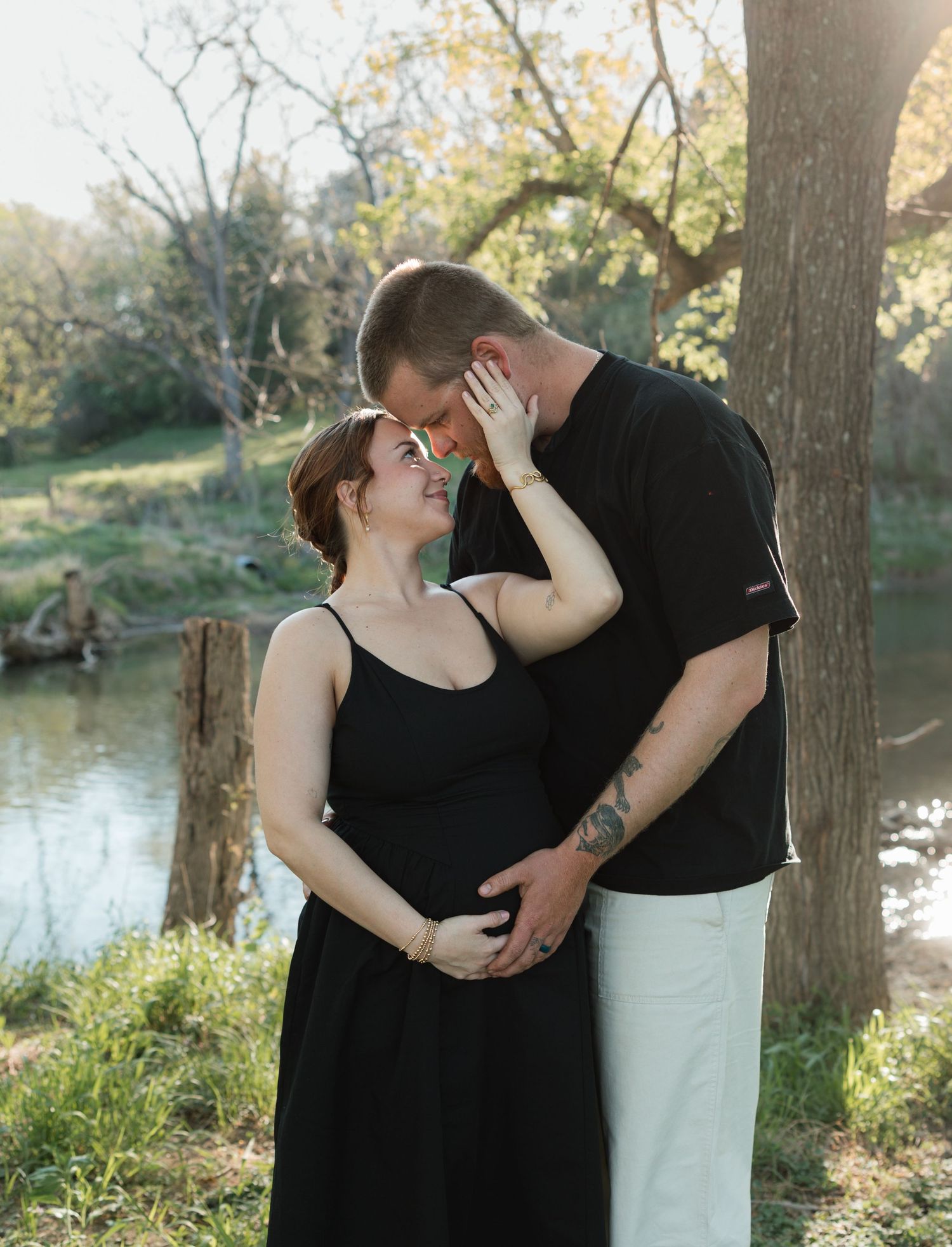Couple sharing a tender moment outdoors by a creek, woman in black dress with baby bump, man in black shirt holding her.