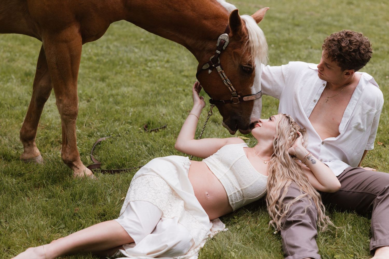 Pregnant woman lying on grass with partner and chestnut horse nuzzling her during romantic maternity photo shoot.