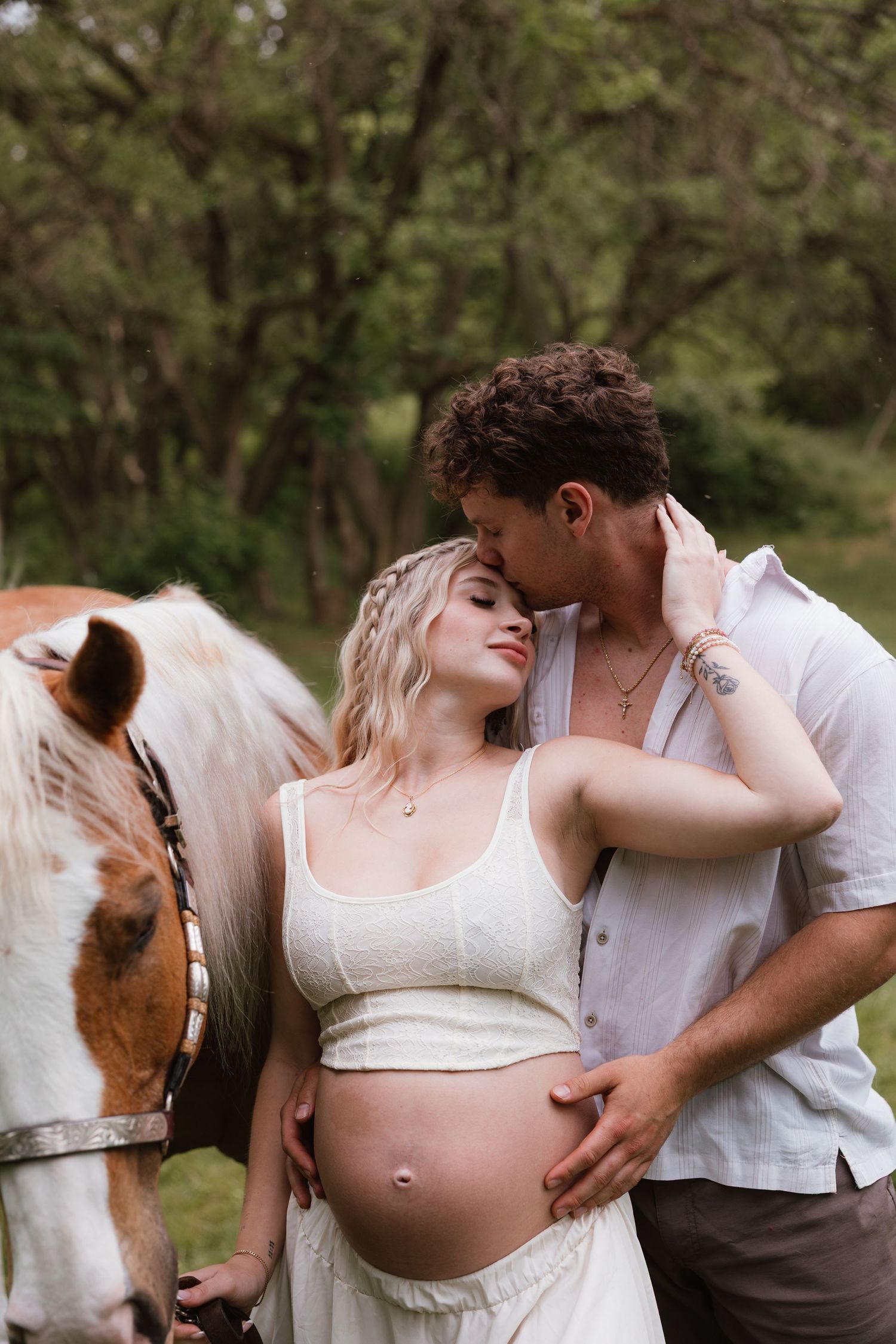 Pregnant woman in white dress with partner kissing her head beside a horse in a lush green outdoor setting.