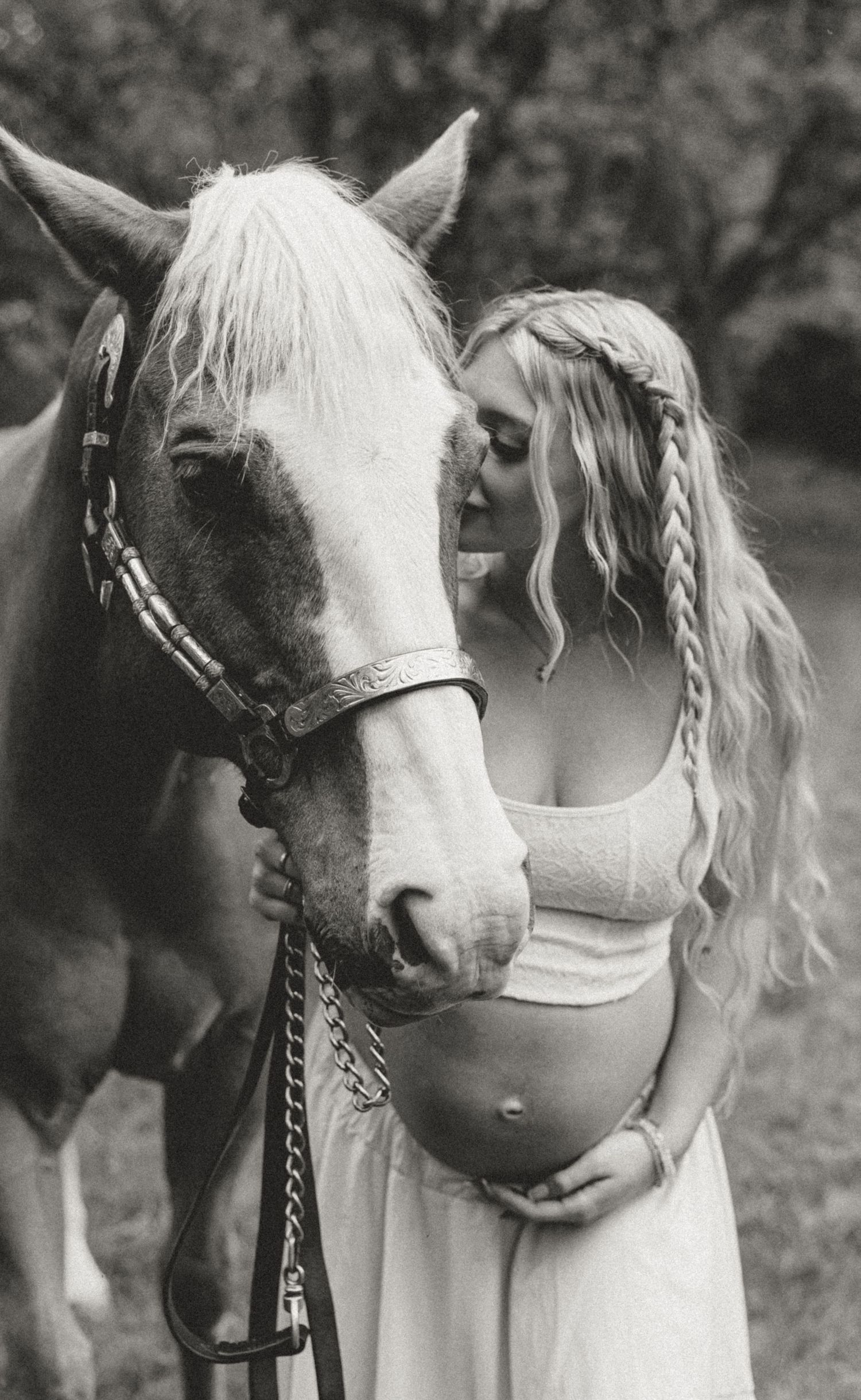 A black and white photo of a pregnant woman in a white dress sharing a tender moment with a white horse in nature.