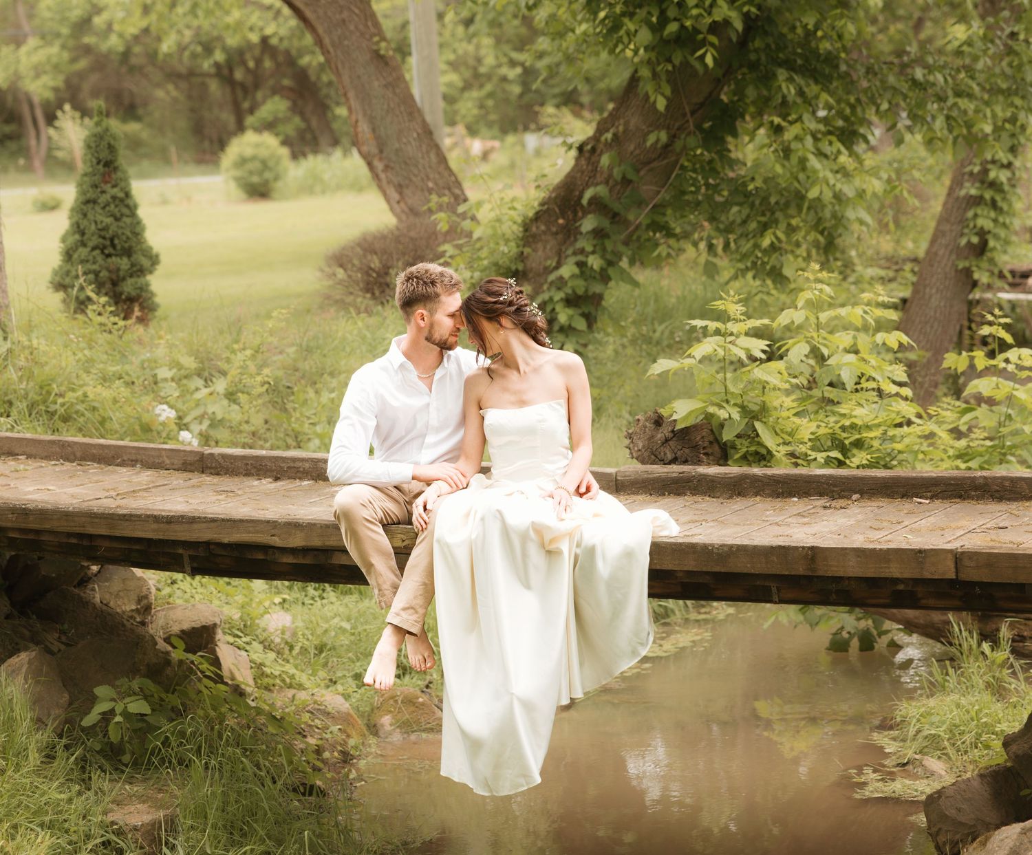 A couple in wedding attire sits intimately on a wooden bridge over a pond in a lush garden setting.