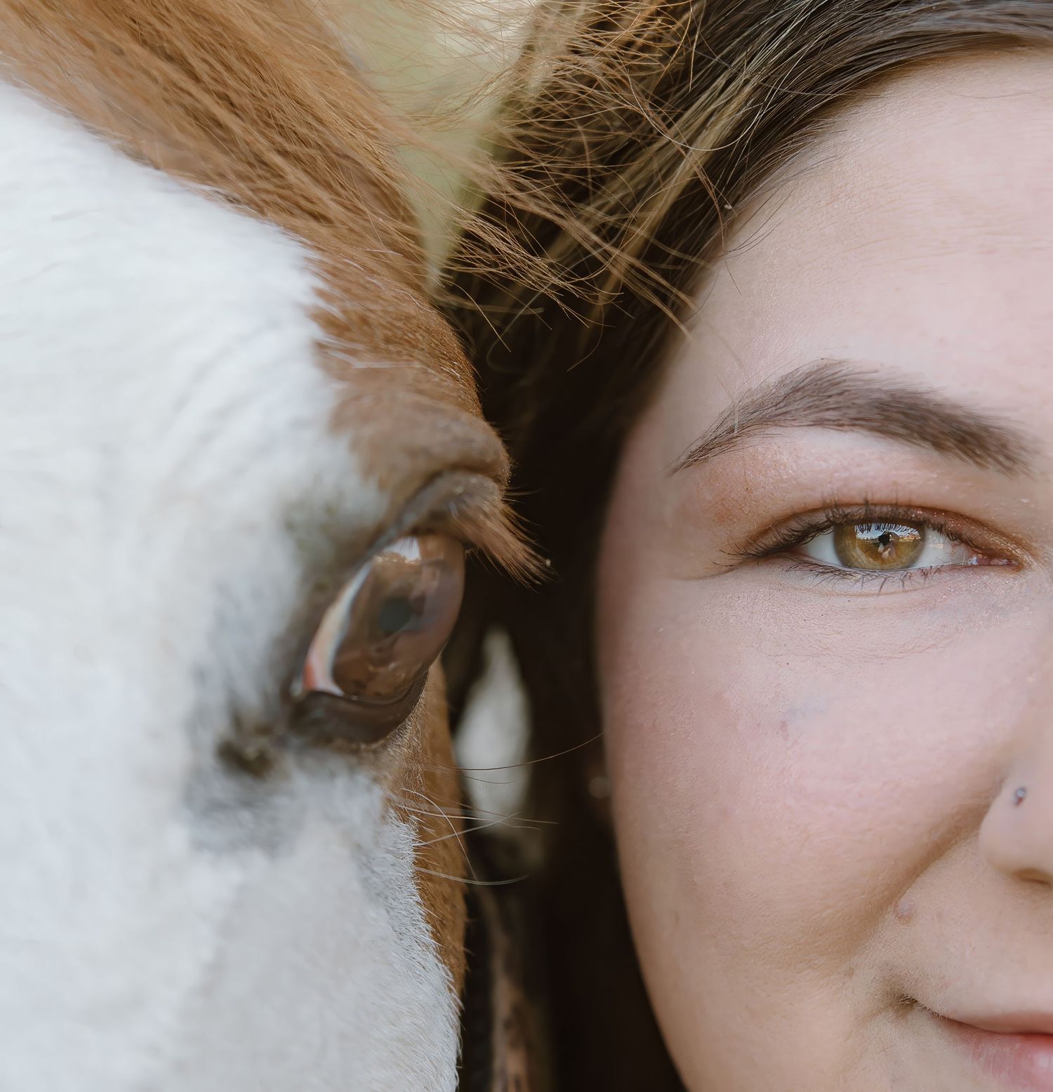 Close-up portrait of a woman with green eyes beside a horse, their faces side by side in an intimate moment.