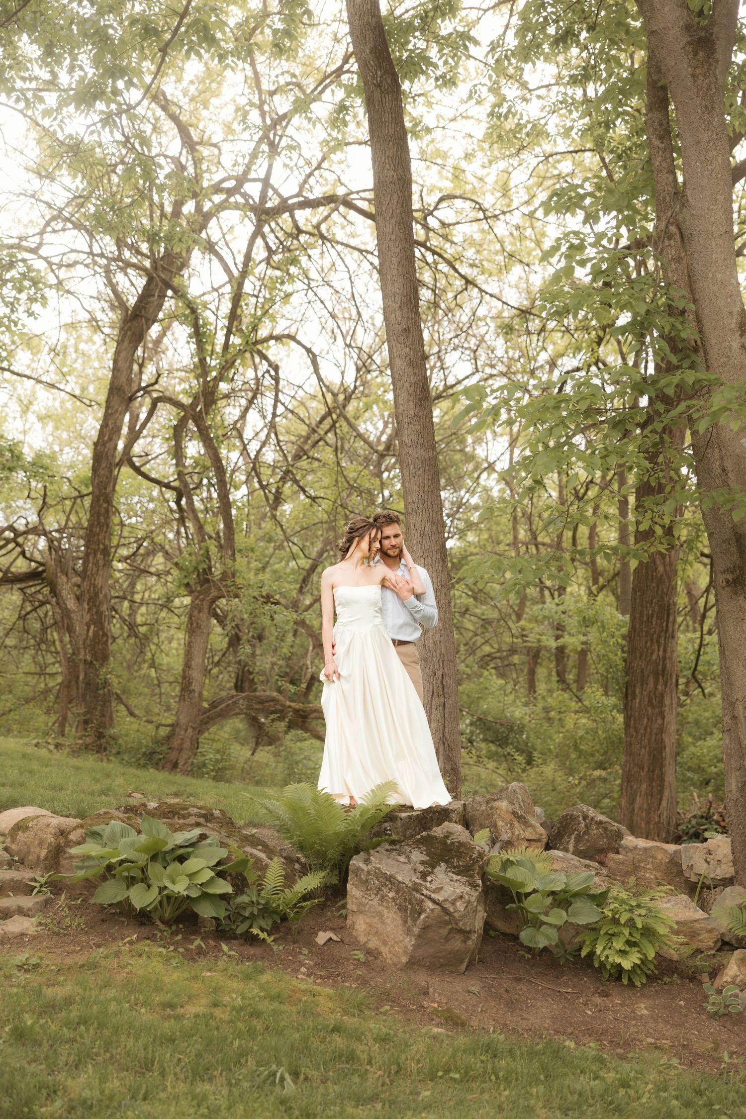 Bride and groom standing on rocks in a lush green forest, bride wearing a flowing white dress.