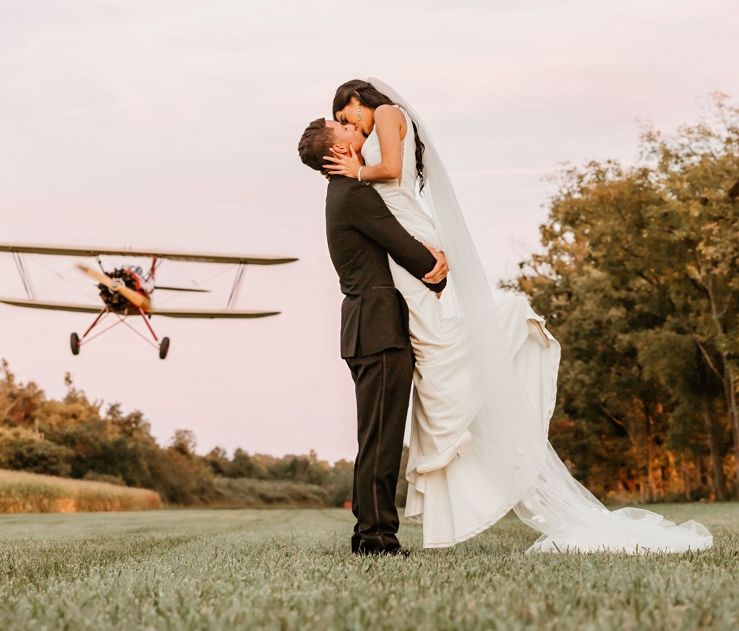 A romantic wedding photo with a vintage airplane flying in the background at sunset.
