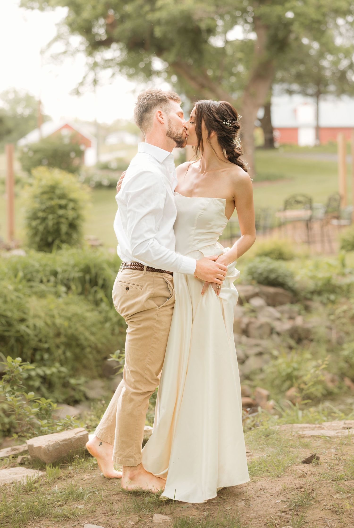 A couple shares a romantic kiss in a lush garden, bride in strapless white gown and groom in casual khaki pants and white shirt.