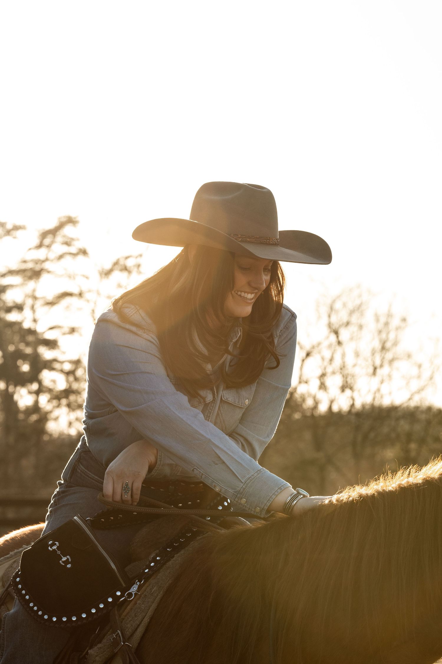A person wearing a cowboy hat rides horseback in warm sunset lighting with a studded leather western saddle.