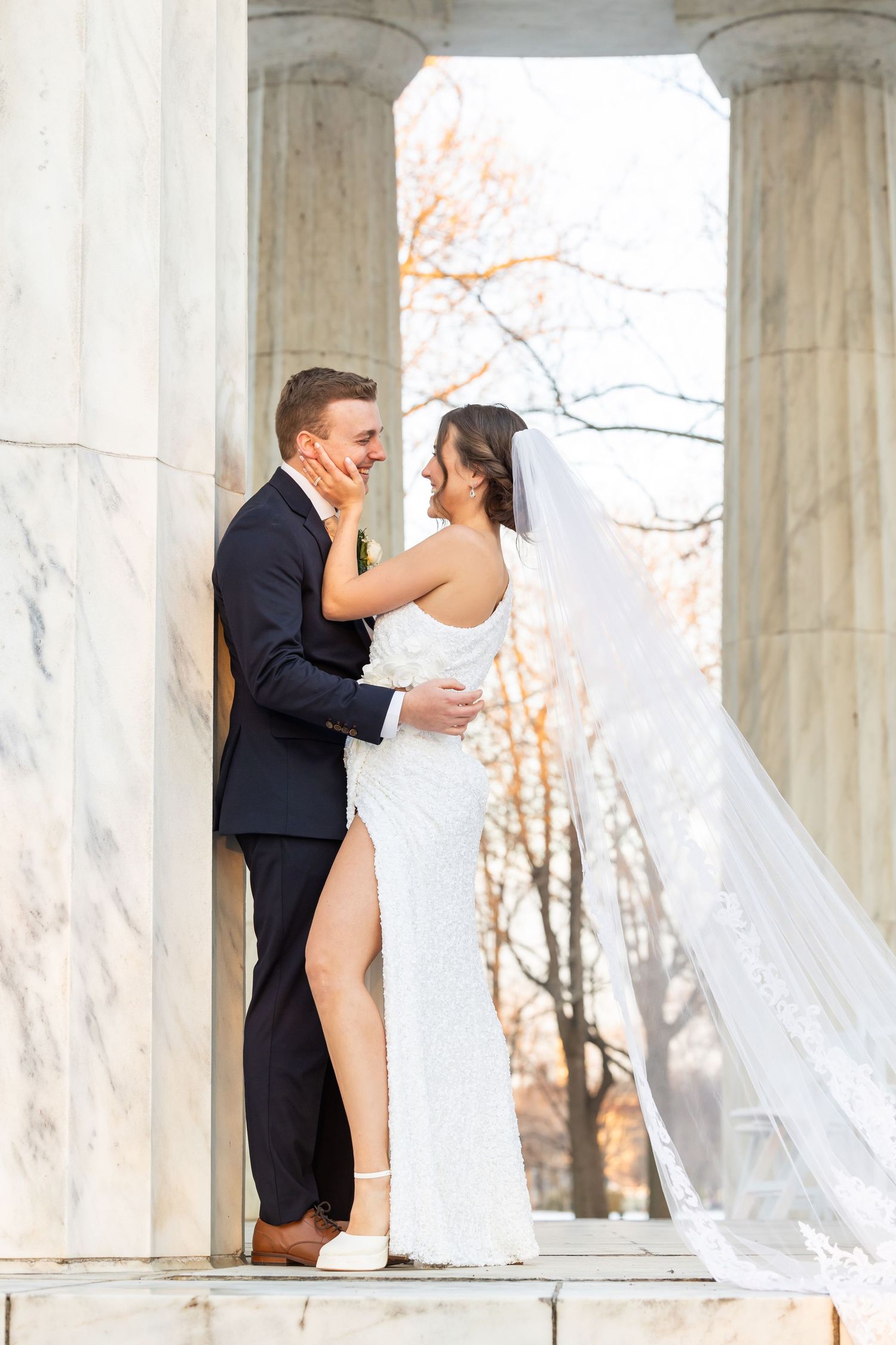 Wedding couple shares intimate moment between classical stone columns with flowing veil in sunlight.