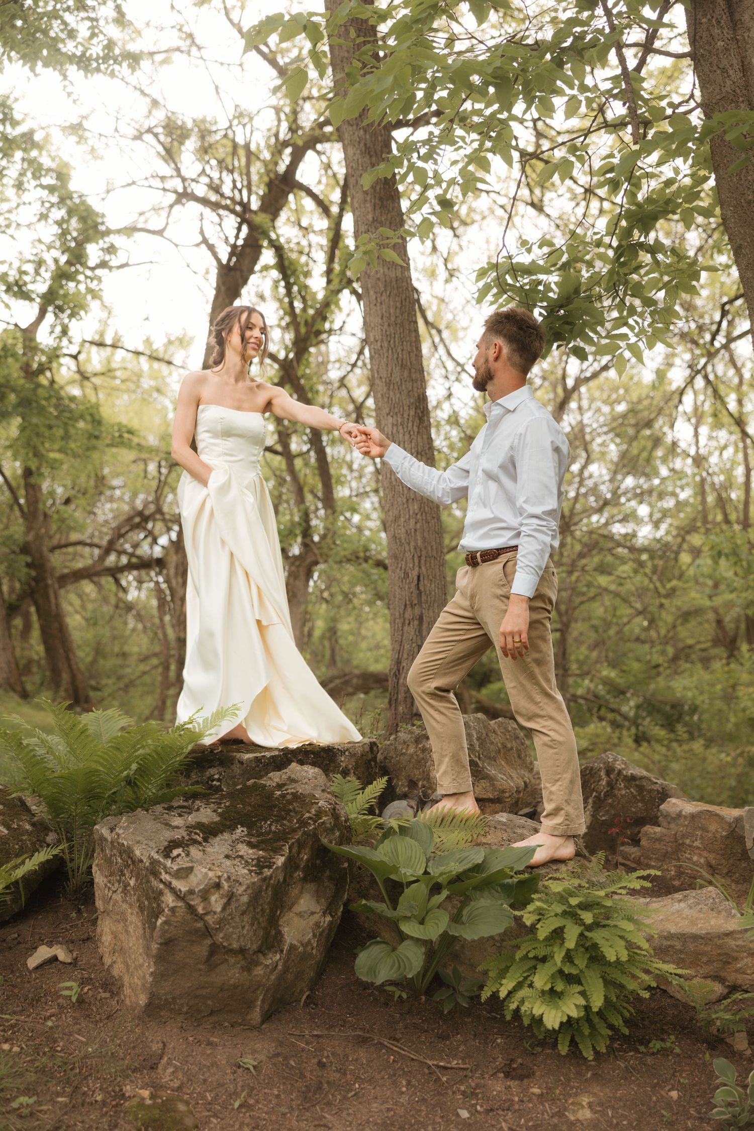 Bride in white gown holds groom's hand on forest rocks during romantic outdoor wedding photoshoot.