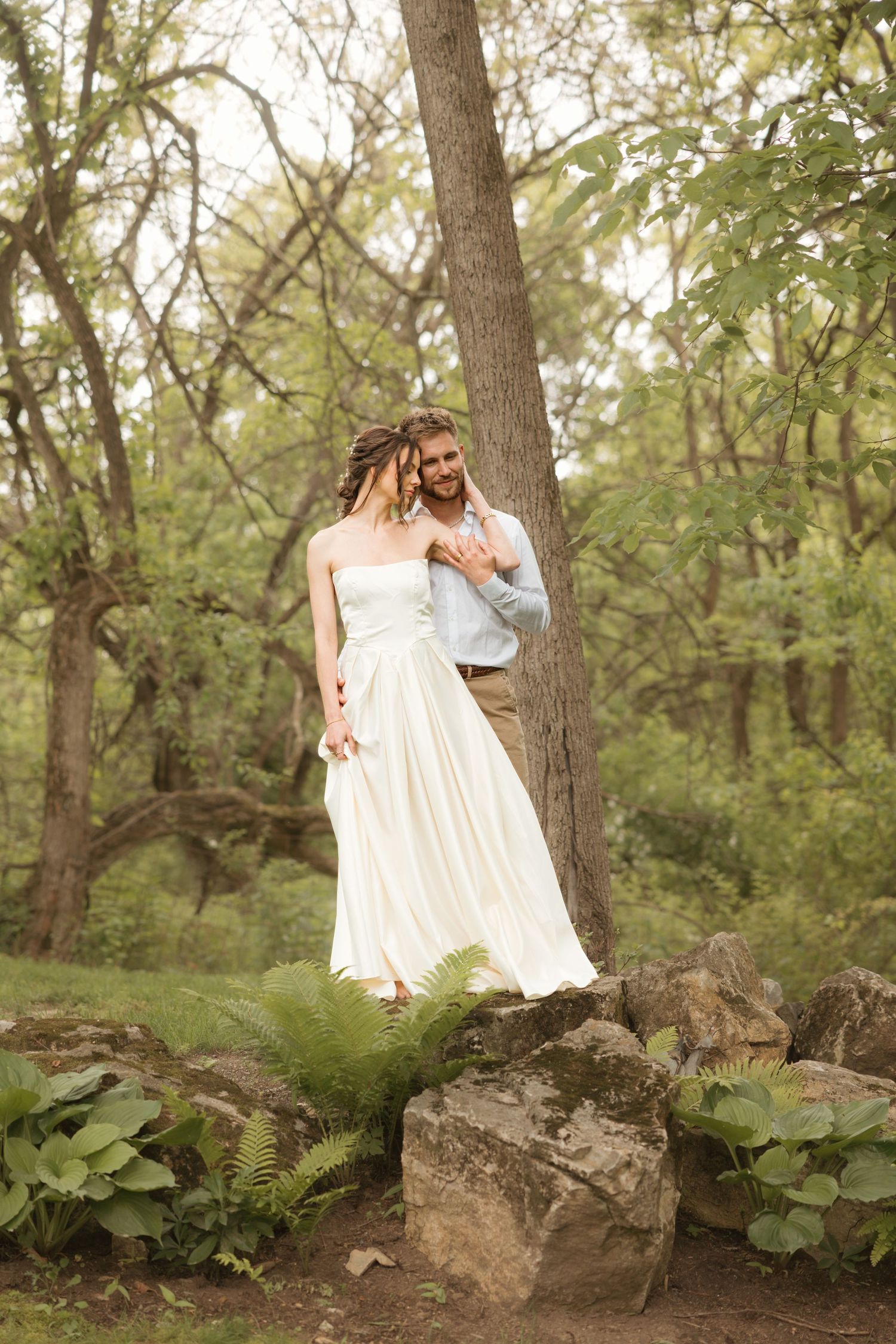 A romantic couple in white outfits share tender moments among trees and ferns in a dreamy woodland setting.