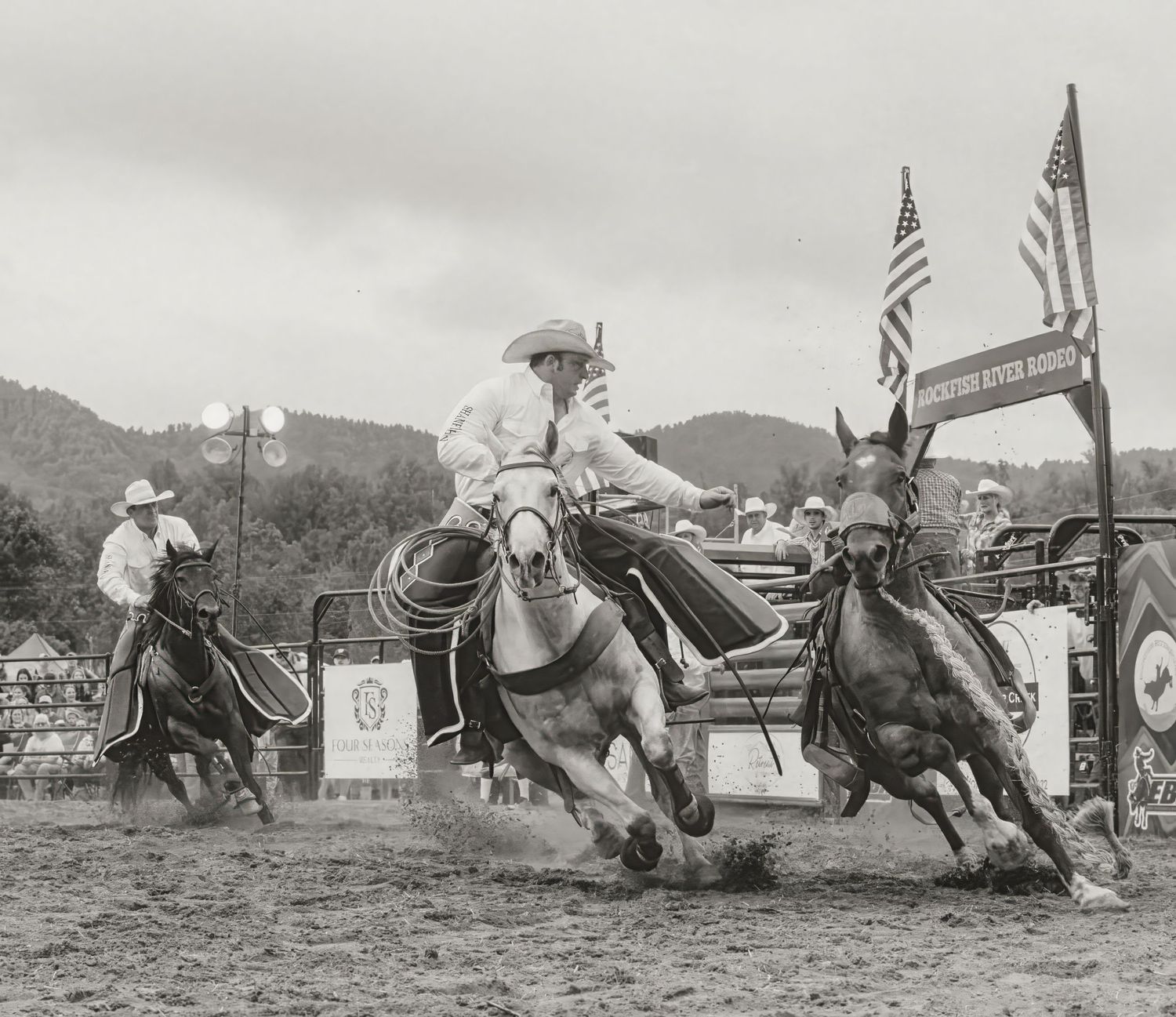 Cowboys compete in a rodeo event with American flags flying as spectators watch from grandstands in the background.