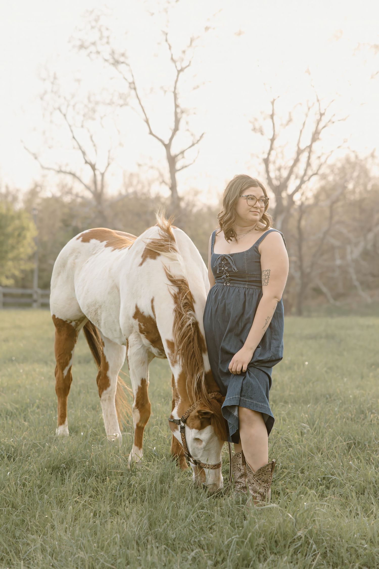Woman in blue dress and cowboy boots stands beside a brown and white paint horse grazing in a sunlit field.