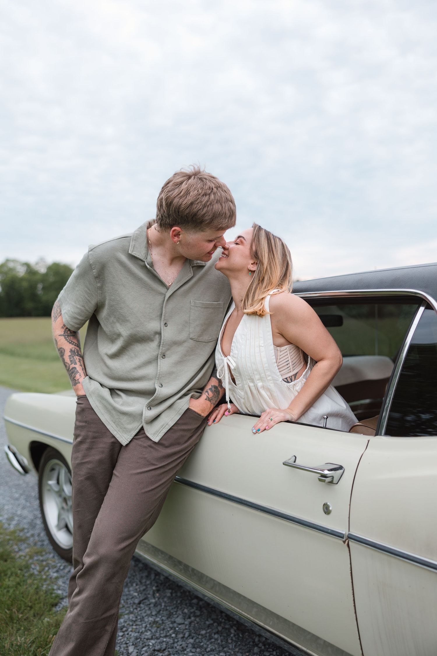 A romantic couple embraces beside a vintage cream-colored car in a countryside setting.