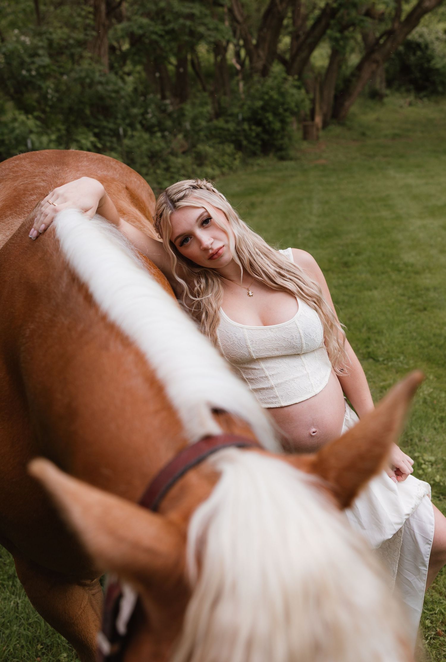Pregnant woman in white crop top leans against a chestnut horse in a lush green outdoor setting.