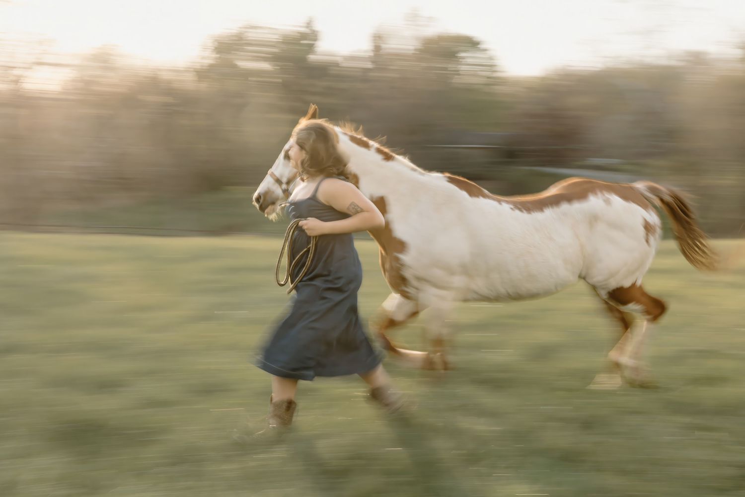 Young woman in a gray dress running with a paint horse across a green field in golden hour light.