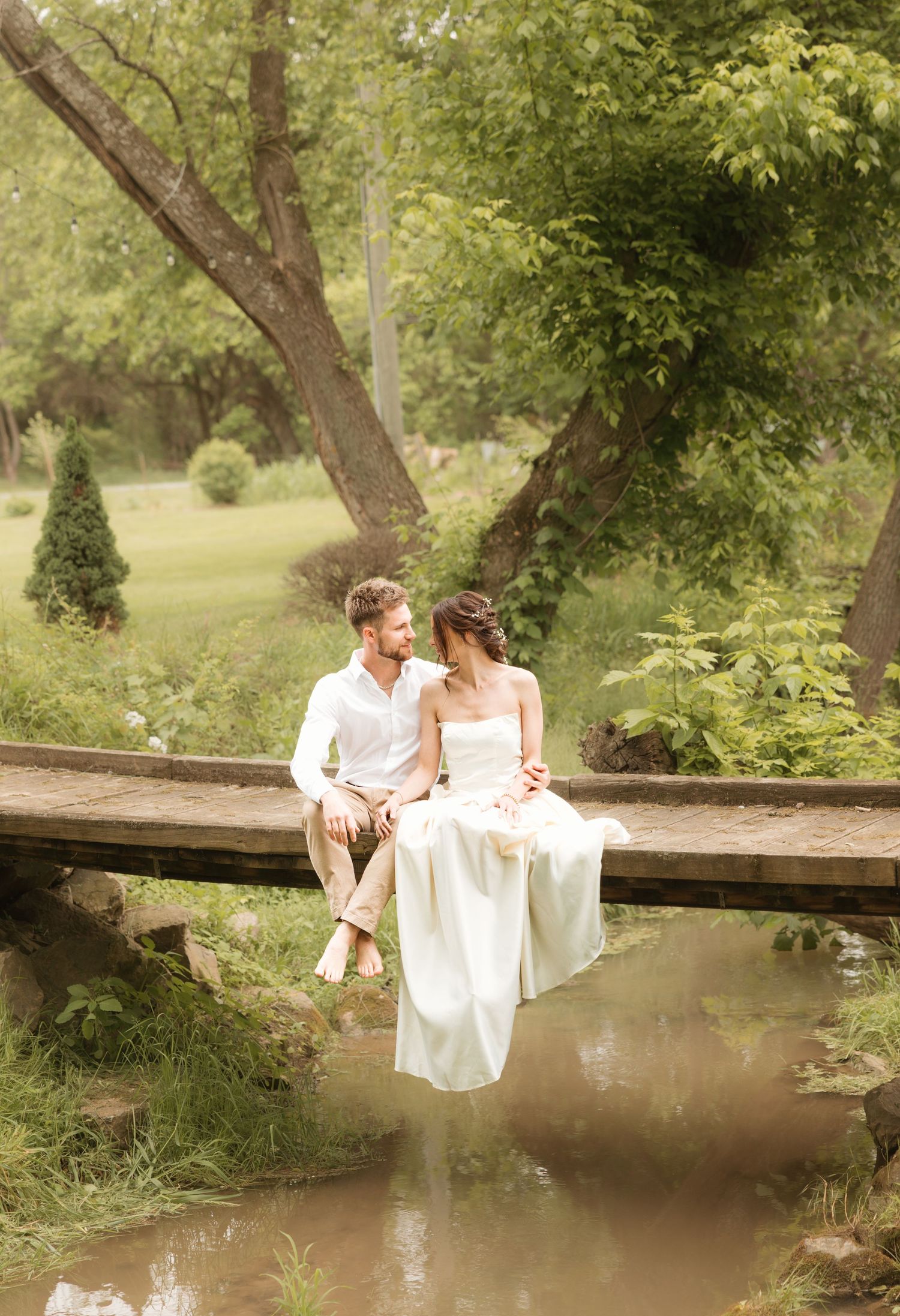 A romantic couple sits together on a wooden footbridge over a tranquil pond in a lush garden setting.