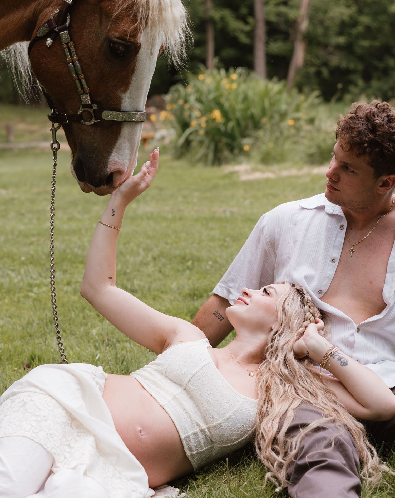 Pregnant woman in white crop top reclines on grass beside a horse while a curly-haired man sits nearby outdoors.