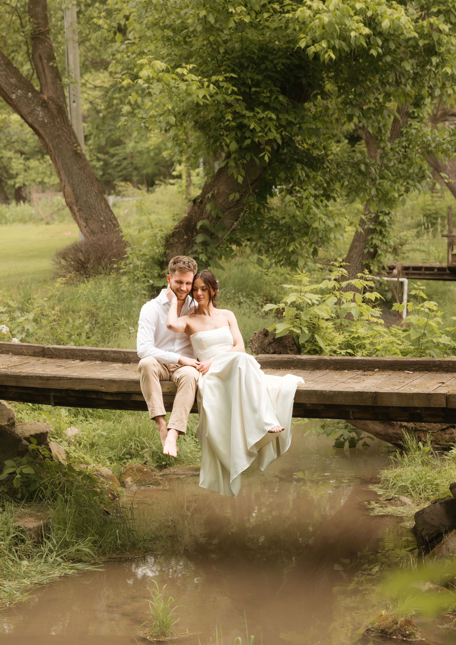 Bride and groom sit together on a wooden bridge over a creek surrounded by lush green trees in an outdoor wedding photo.