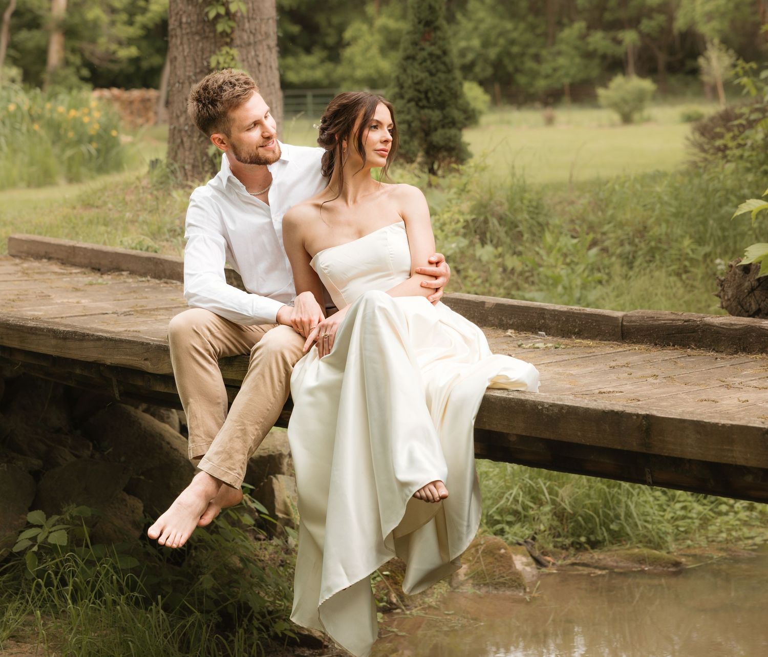 A couple in wedding attire sits together on a rustic wooden bridge in a lush outdoor setting.