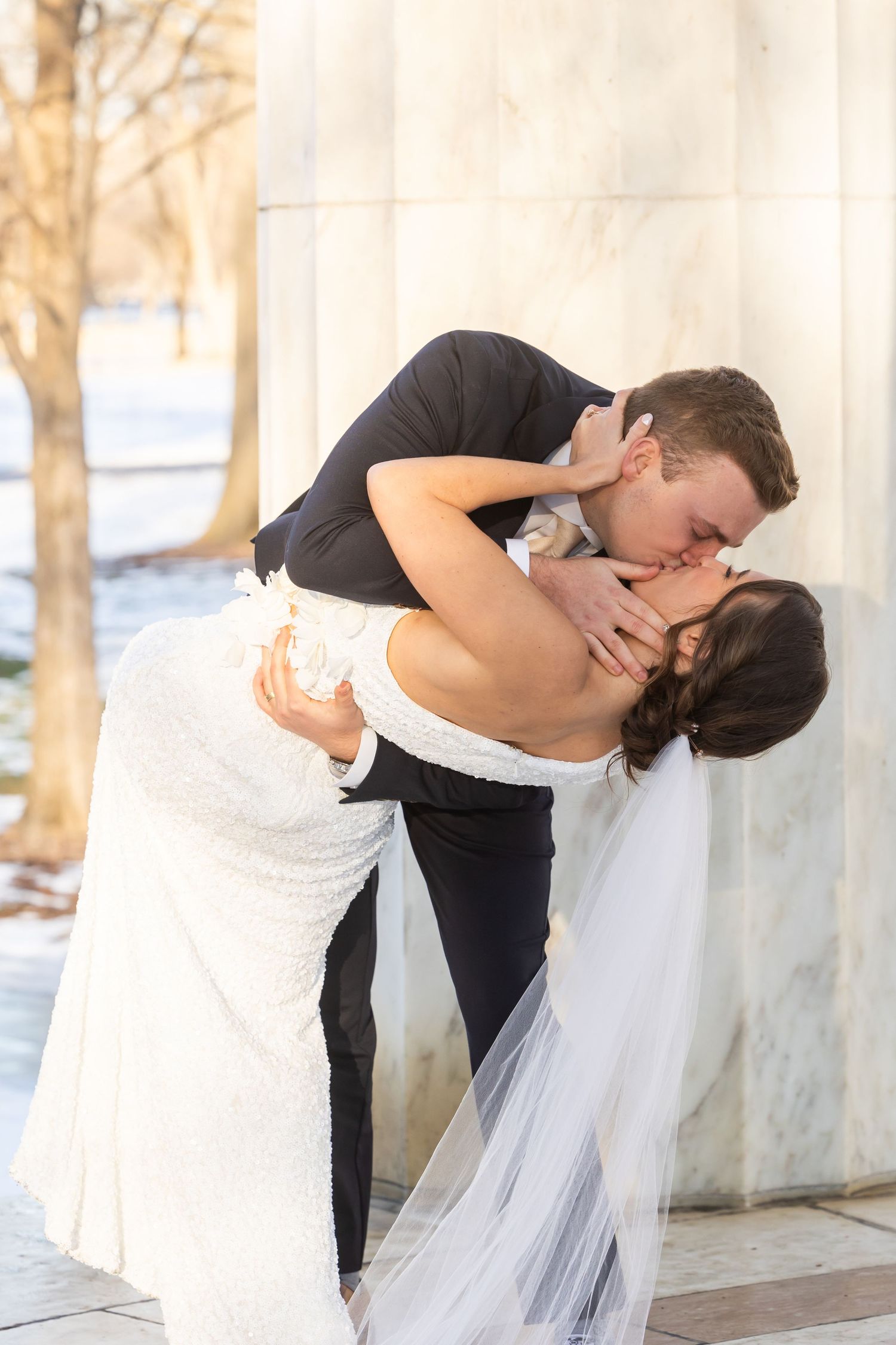 A romantic wedding dip kiss captured in natural light against white marble columns.