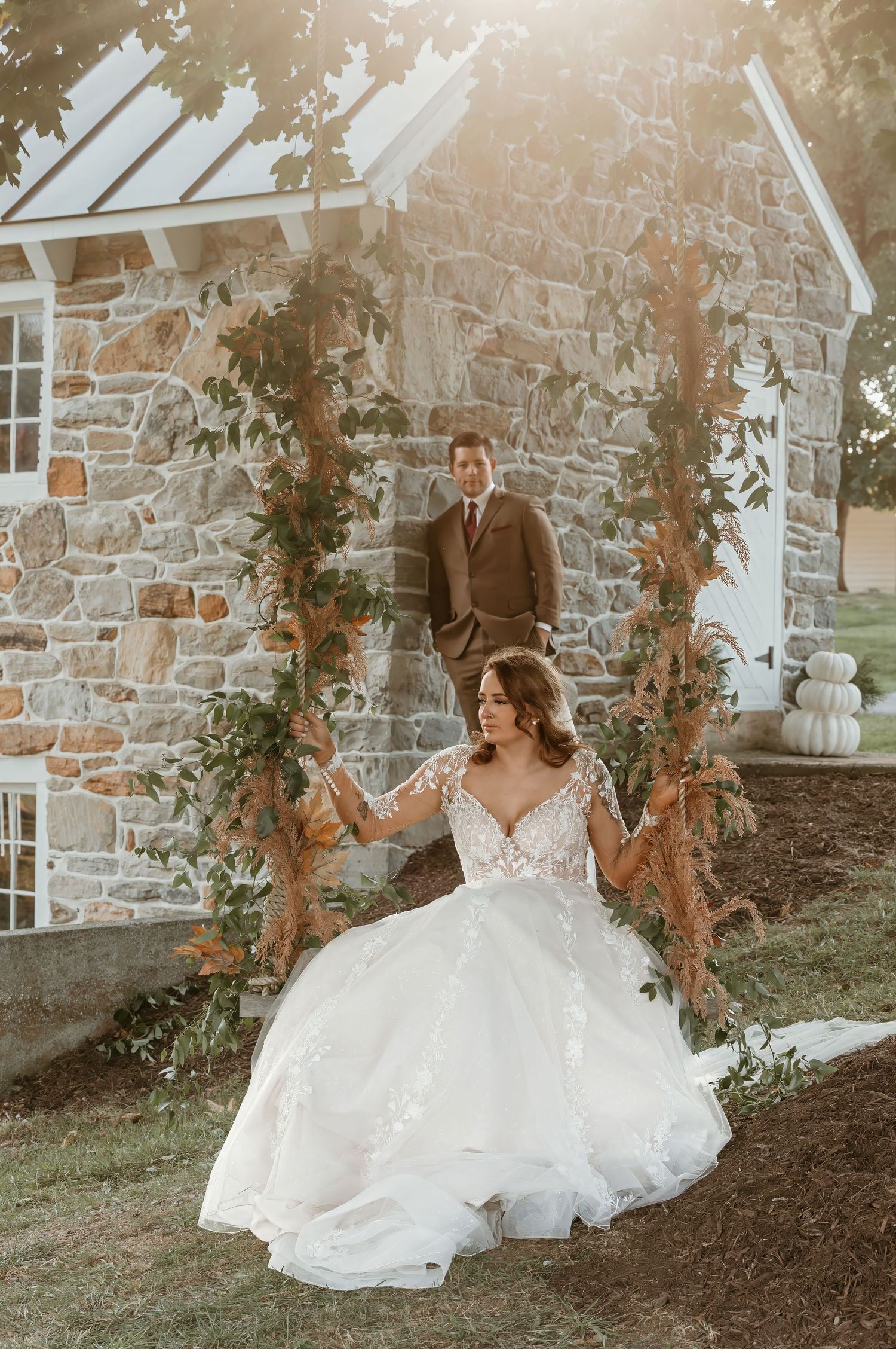 A romantic wedding photo of a bride on a decorative swing against a rustic stone building at sunset.