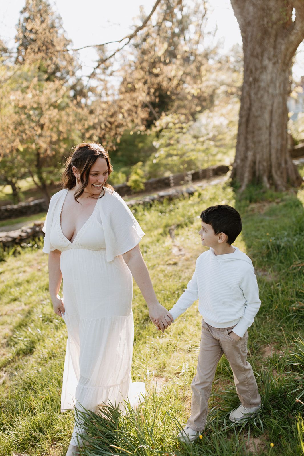 Mother and son hold hands in a sunlit outdoor setting, wearing coordinating white outfits.