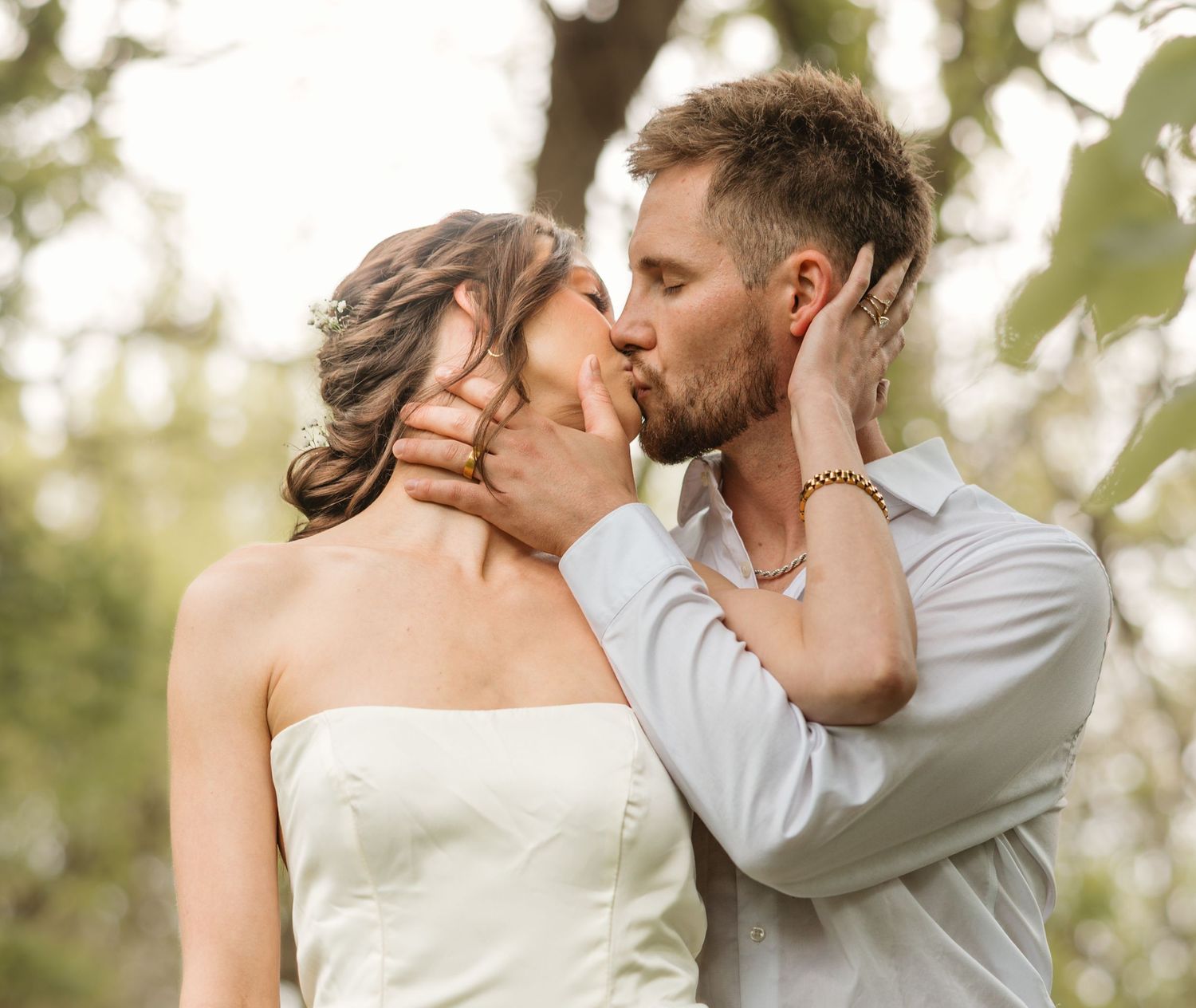 A romantic couple shares a tender kiss outdoors in a natural setting with soft sunlight filtering through trees.