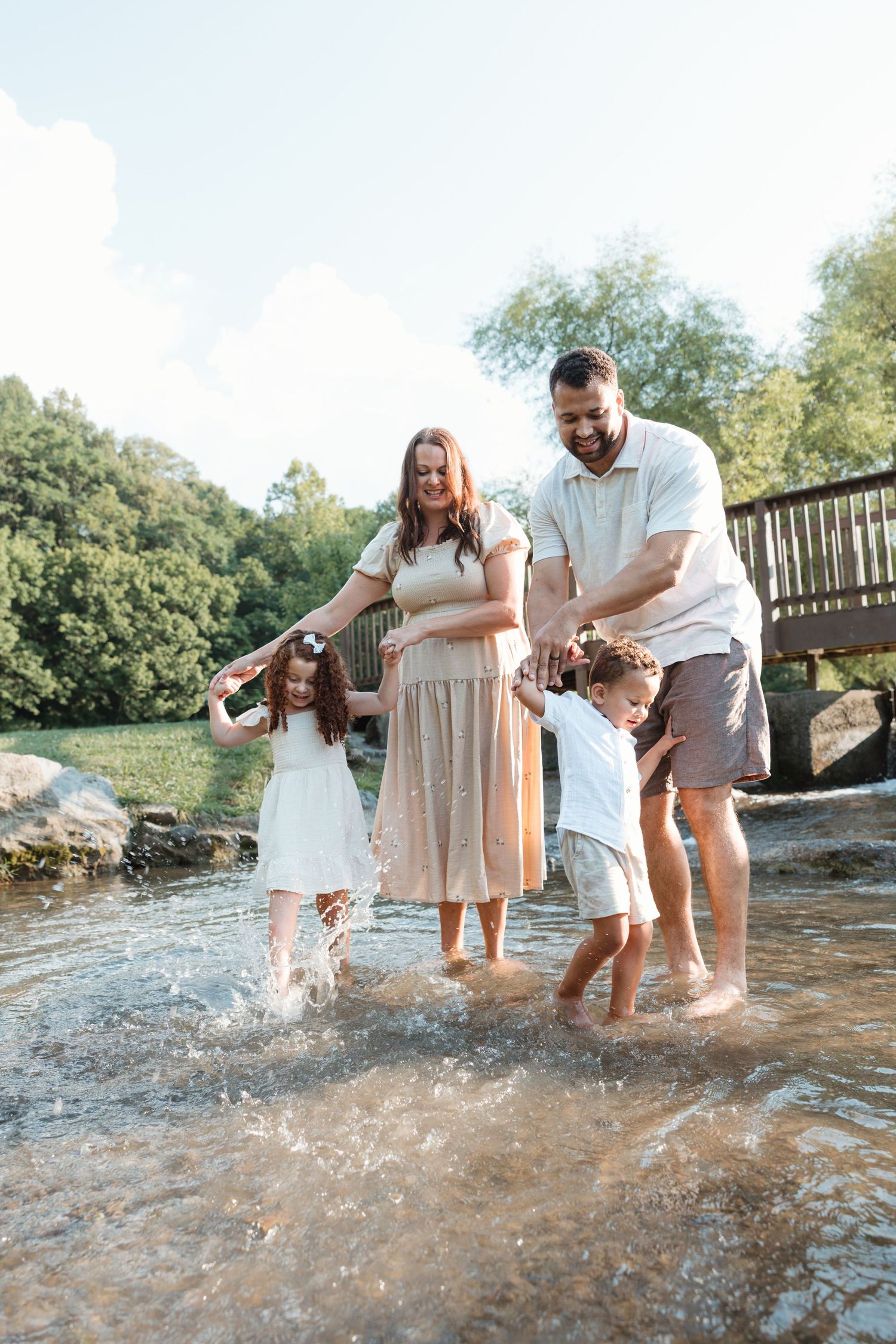 Parents and child playfully splash and laugh together in shallow river water while exploring on a sunny day in nature.