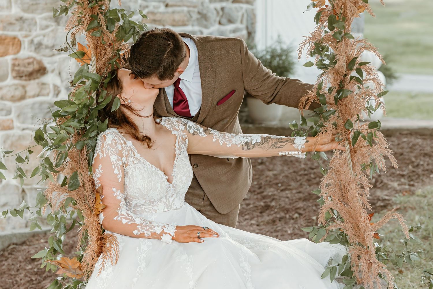 Romantic wedding couple shares kiss on decorated rustic swing with floral accents.