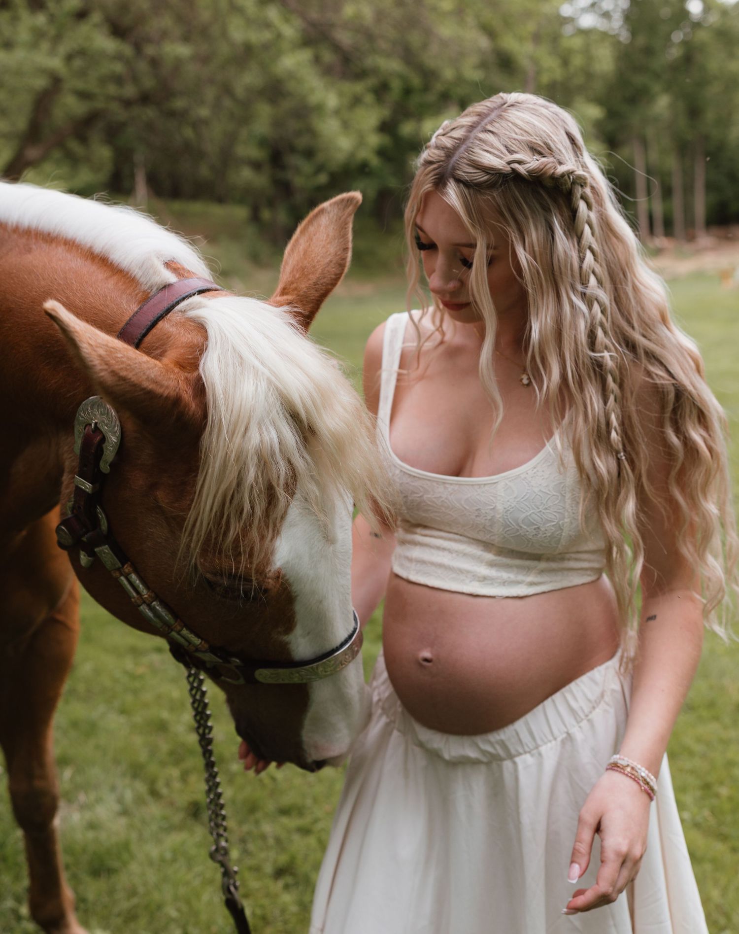 Pregnant woman in white crop top and skirt with braided blonde hair tenderly interacts with a chestnut horse outdoors.