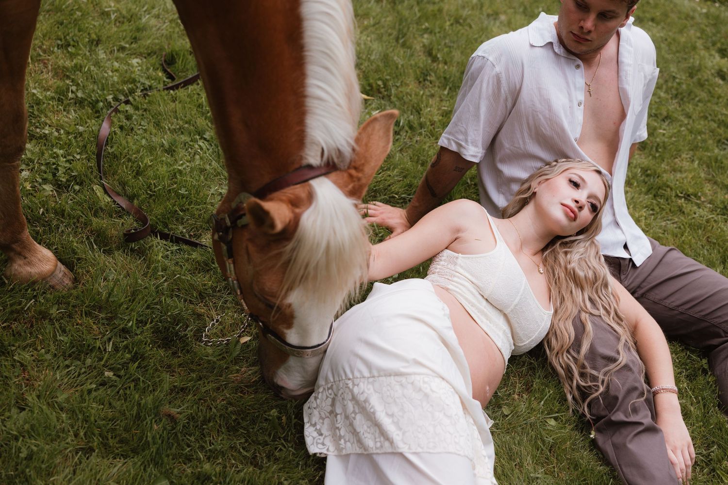 Pregnant woman in white crop top reclines on grass beside a horse while a man sits nearby outdoors.